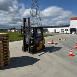 Żółty wózek widłowy Jungheinrich na placu manewrowym, ustawione palety i pachołki, w tle budynek magazynowy i słup wysokiego napięcia. Kurs obsługi wózka.