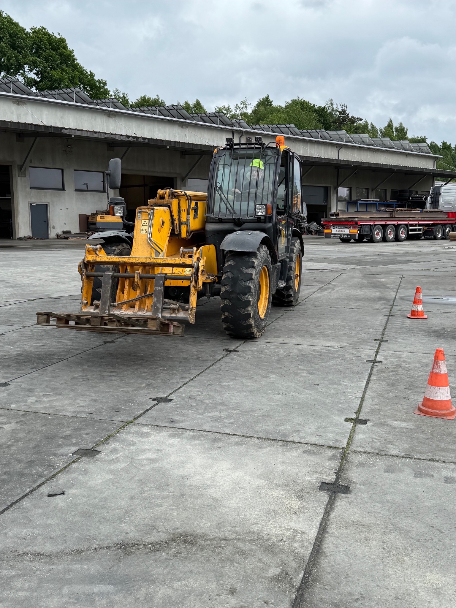 Żółto-czarny wózek widłowy teleskopowy na betonowym placu, w tle budynek magazynowy i ciężarówka z ładunkiem. Dwa pachołki ostrzegawcze z przodu.