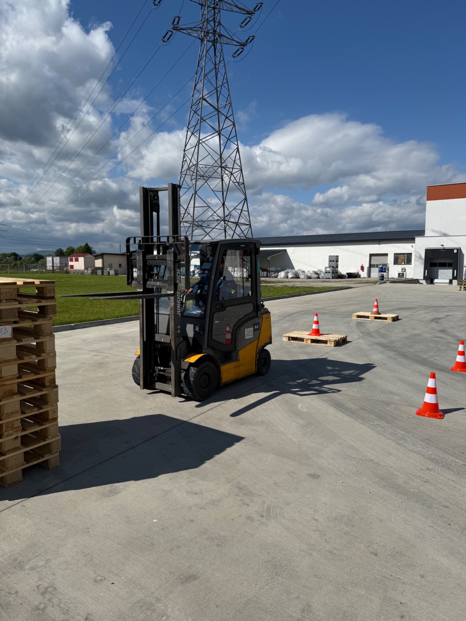 Żółty wózek widłowy Jungheinrich na placu manewrowym, ustawione palety i pachołki, w tle budynek magazynowy i słup wysokiego napięcia. Kurs obsługi wózka.