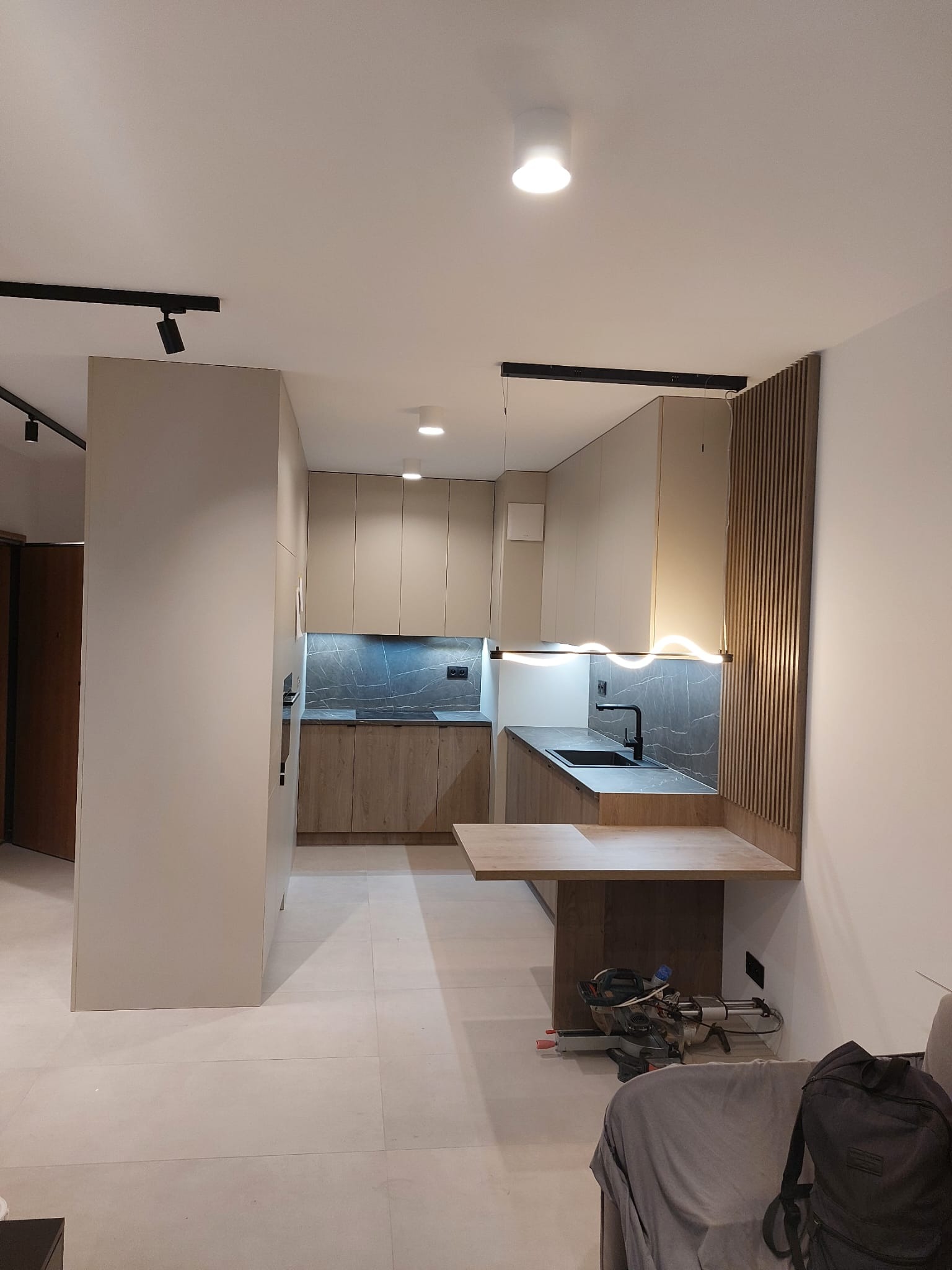 Nowoczesna kuchnia z drewnianymi i beżowymi frontami, czarnym blatem i oświetleniem LED. Widoczne narzędzia sugerują etap montażu.