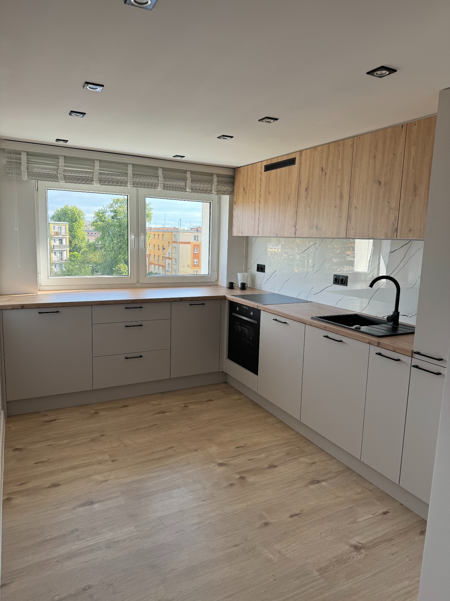 Jasna kuchnia z drewnianymi akcentami: szare szafki dolne, drewniane górne, marmurowy backsplash, czarna armatura i okno z widokiem na zieleń miejską.