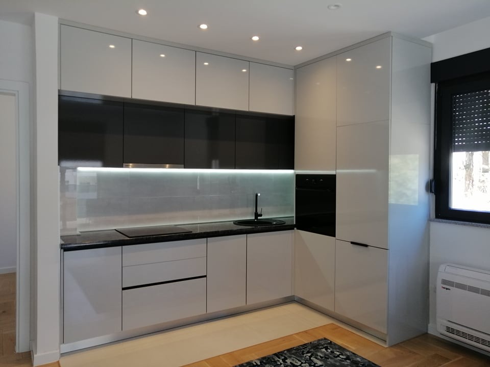Nowoczesna kuchnia na wymiar z białymi, lakierowanymi frontami i czarnymi akcentami. Widoczny zlew, płyta indukcyjna i oświetlenie LED pod szafkami.