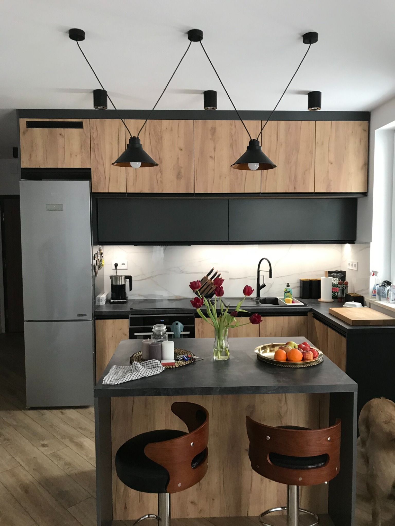 Nowoczesna kuchnia z drewnianymi szafkami, czarnymi blatami i wyspą. Nad wyspą wiszą lampy w stylu industrialnym. Na blacie owoce i kwiaty.