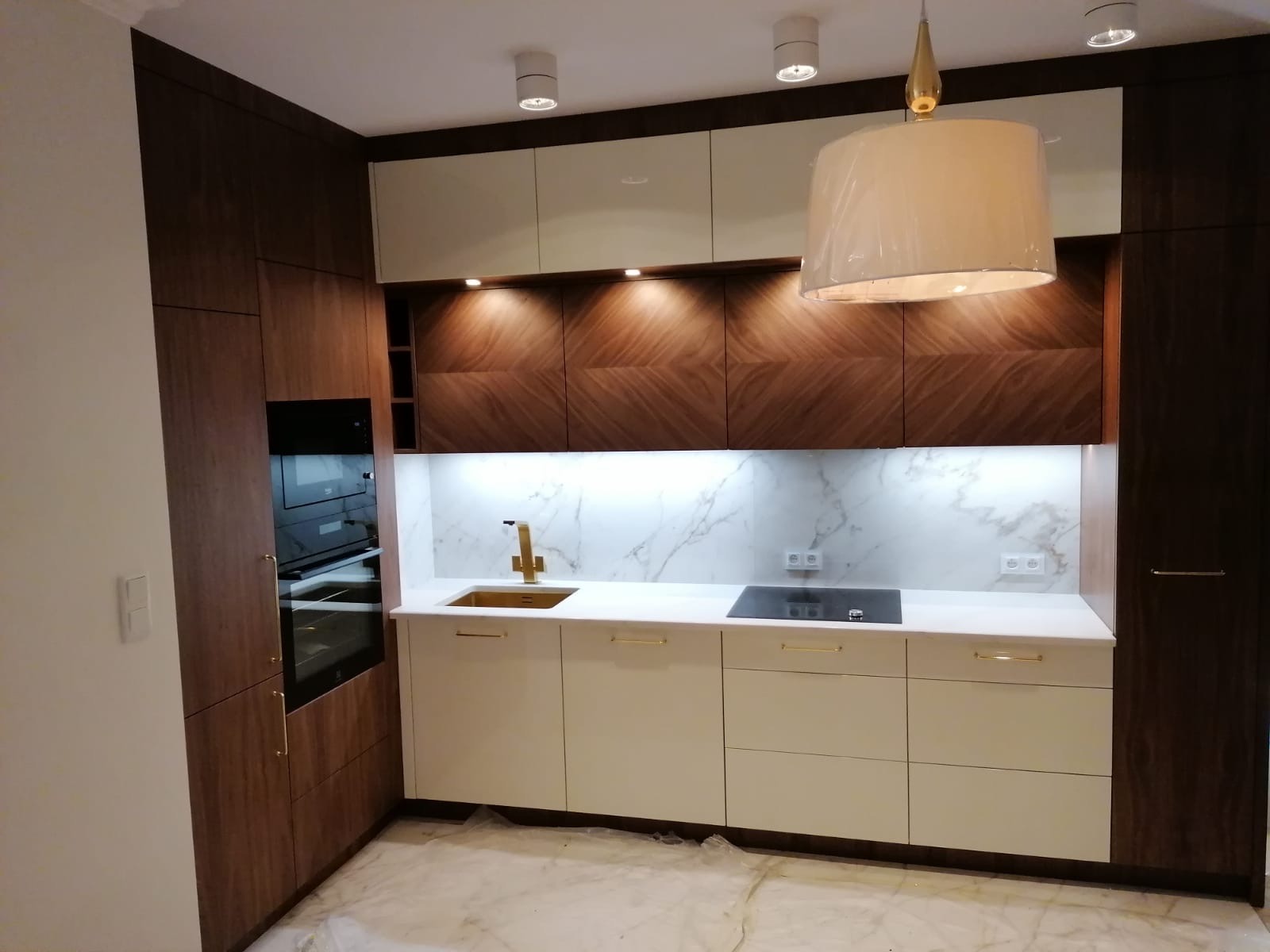 Nowoczesna kuchnia z drewnianymi szafkami, białym blatem i złotą armaturą, oświetlona lampą wiszącą, prezentuje eleganckie wykończenie wnętrza.