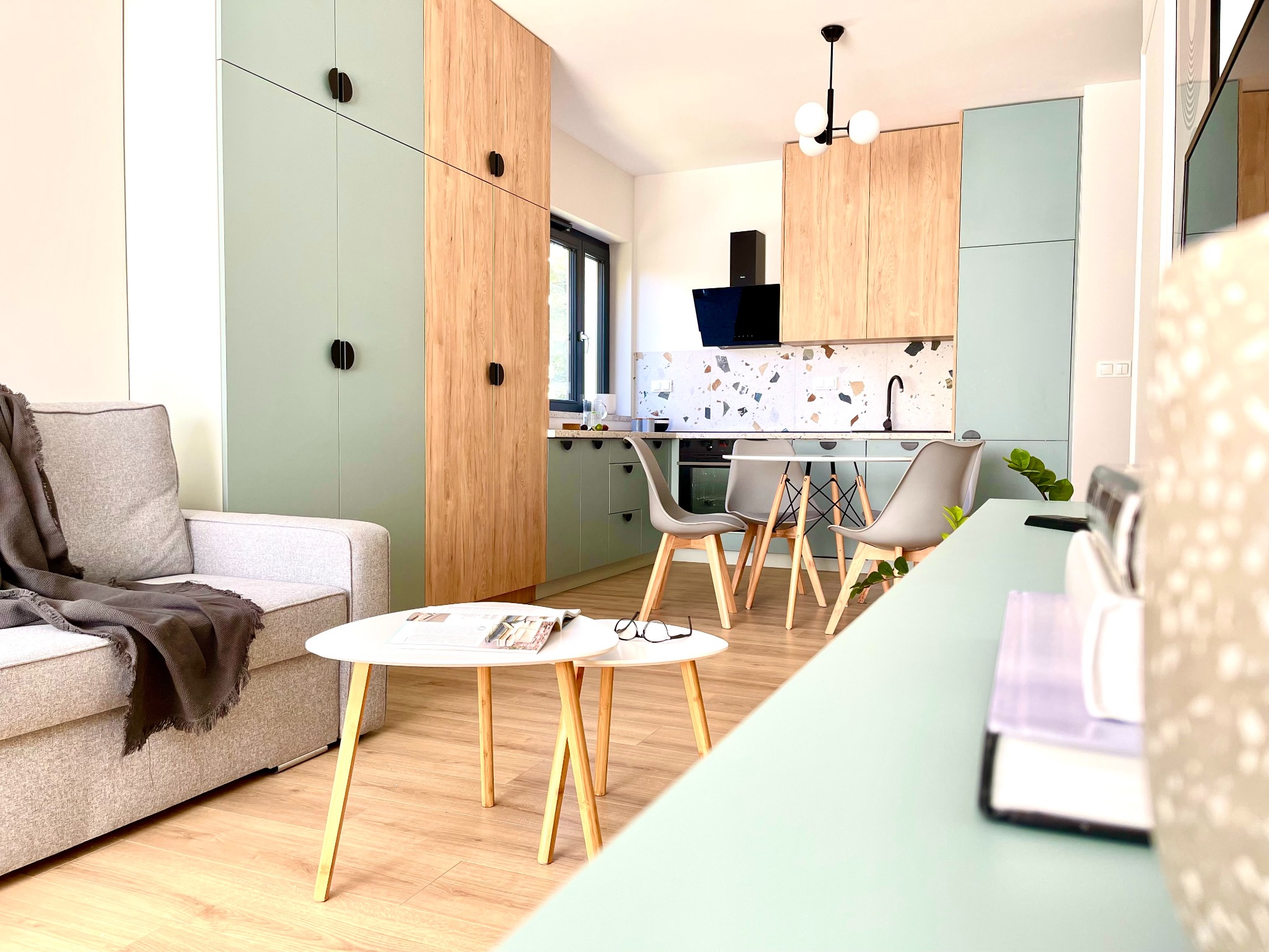 Jasna kuchnia z drewnianymi i miętowo-zielonymi frontami szafek, okrągły stół z krzesłami i szara sofa z kocem w tle. Nowoczesny wystrój wnętrza.