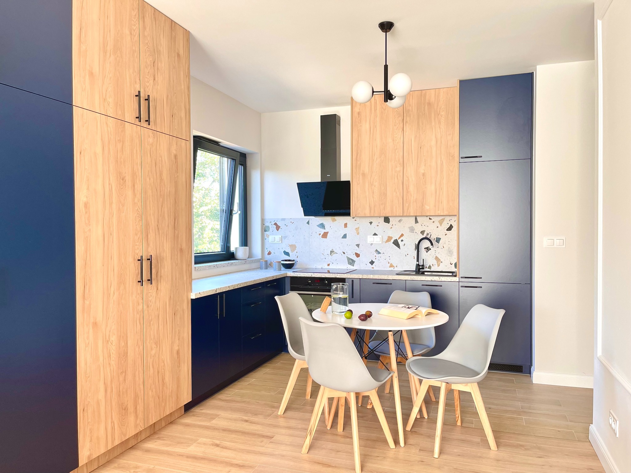 Nowoczesna kuchnia z drewnianymi i granatowymi szafkami, okrągłym stołem i wzorzystą ścianą nad blatem. Minimalistyczny design i jasne oświetlenie.