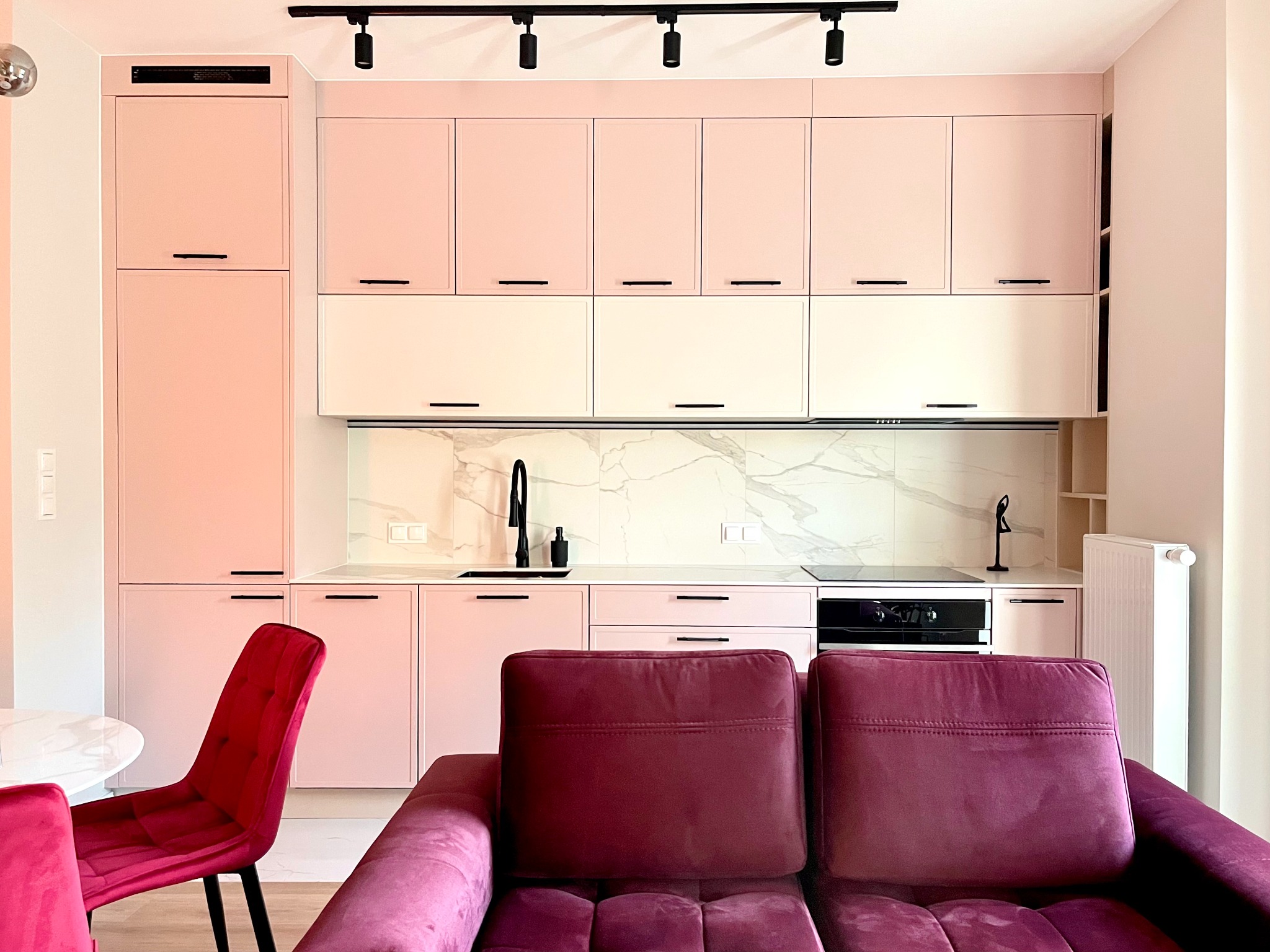 Różowa kuchnia z czarnymi akcentami: armatura, uchwyty i oświetlenie. Marmurowy backsplash i bordowa sofa w kadrze. Nowoczesne i eleganckie wykończenie wnętrza.