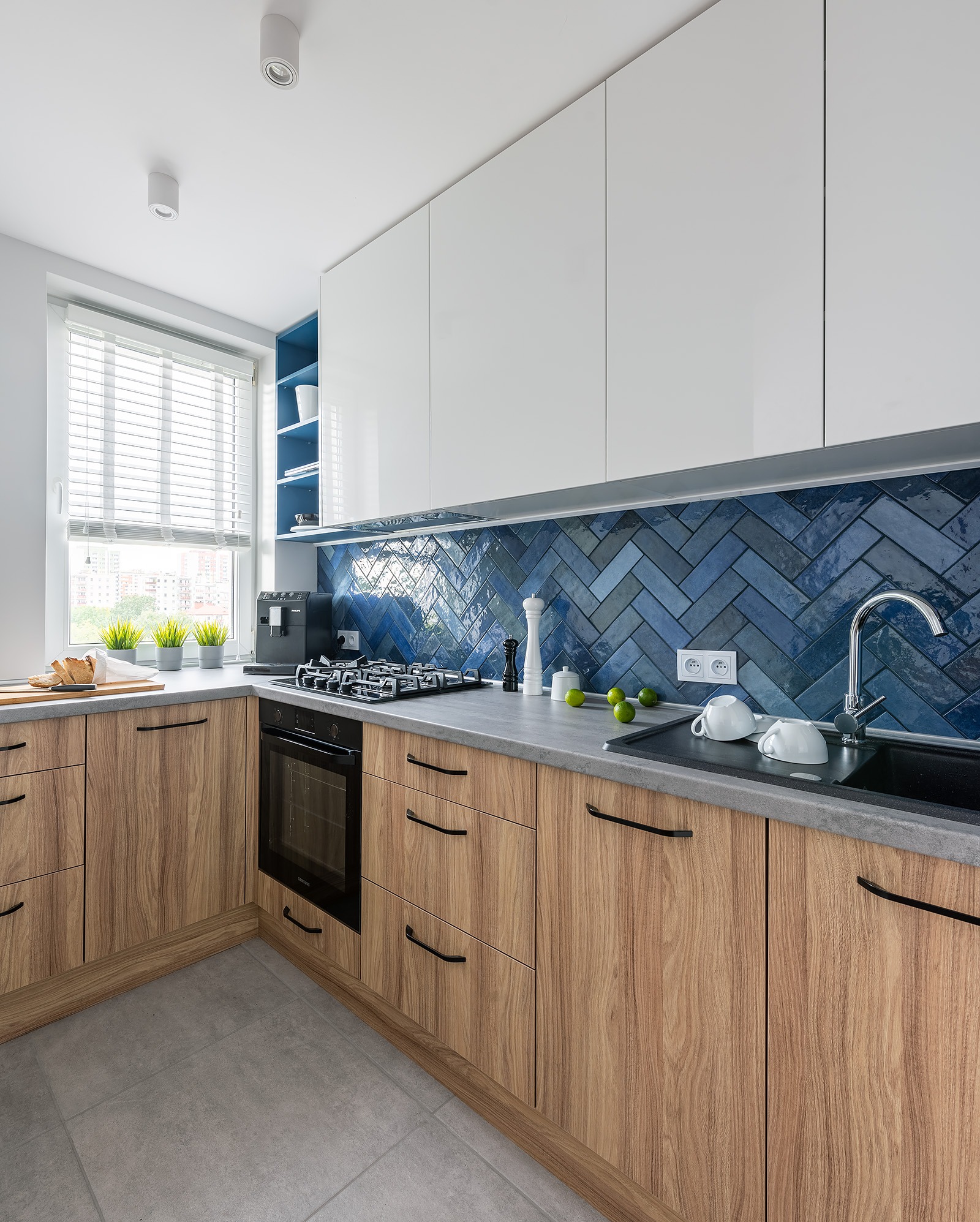 Nowoczesna kuchnia z drewnianymi szafkami, białymi górnymi frontami i niebieską mozaiką ułożoną w jodełkę nad blatem. Widok na okno z żaluzjami.