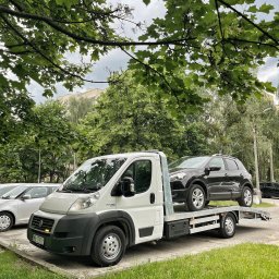Carrier Cars Oskar Konior - Laweta z czarnym SUV-em na platformie, zaparkowana na parkingu w otoczeniu zieleni miejskiej. Widoczne inne zaparkowane auta i budynek w tle.