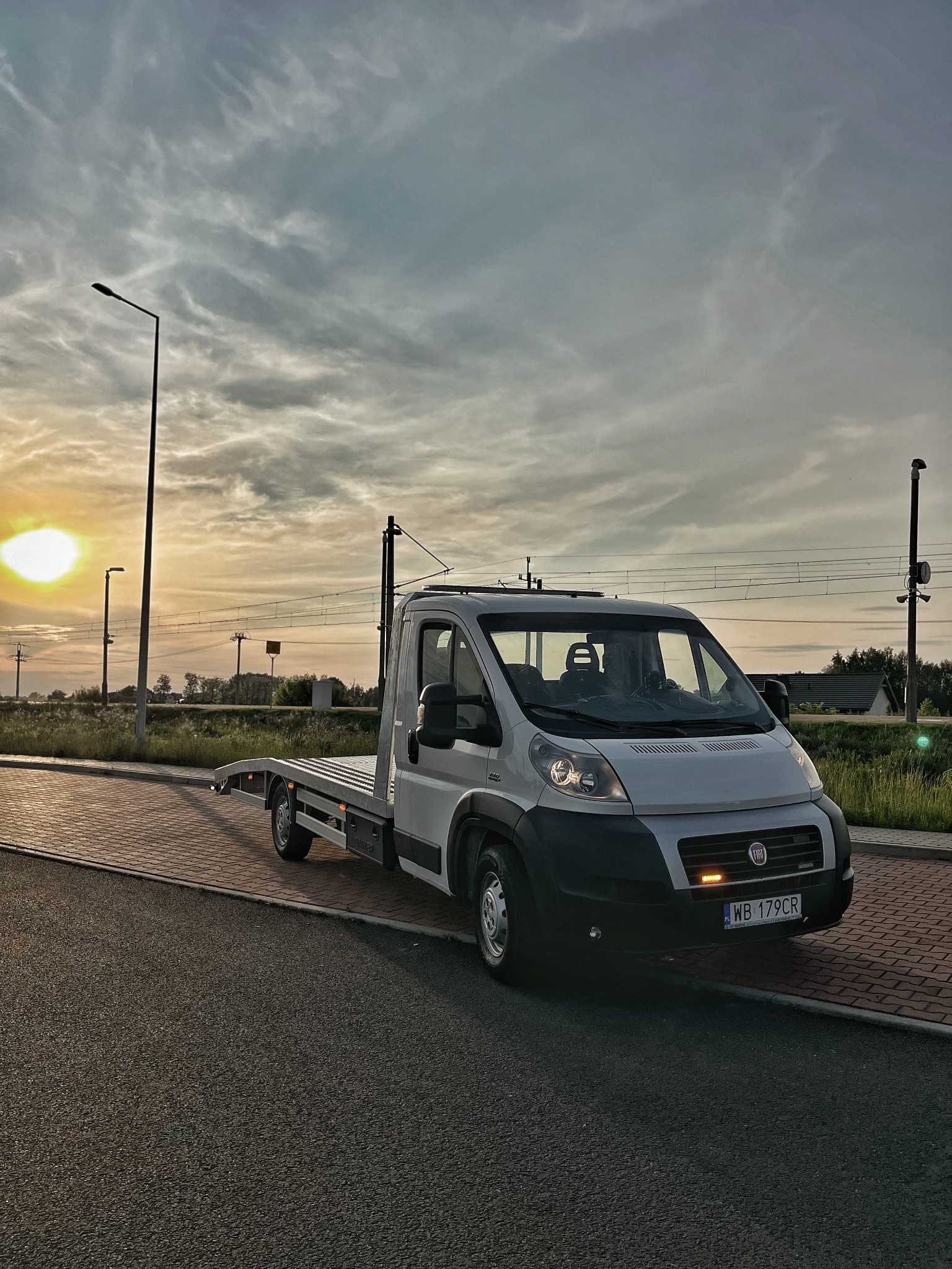 Biała laweta Fiat na tle zachodzącego słońca, zaparkowana na poboczu z kostki brukowej. Widoczne tablice rejestracyjne i światła awaryjne.