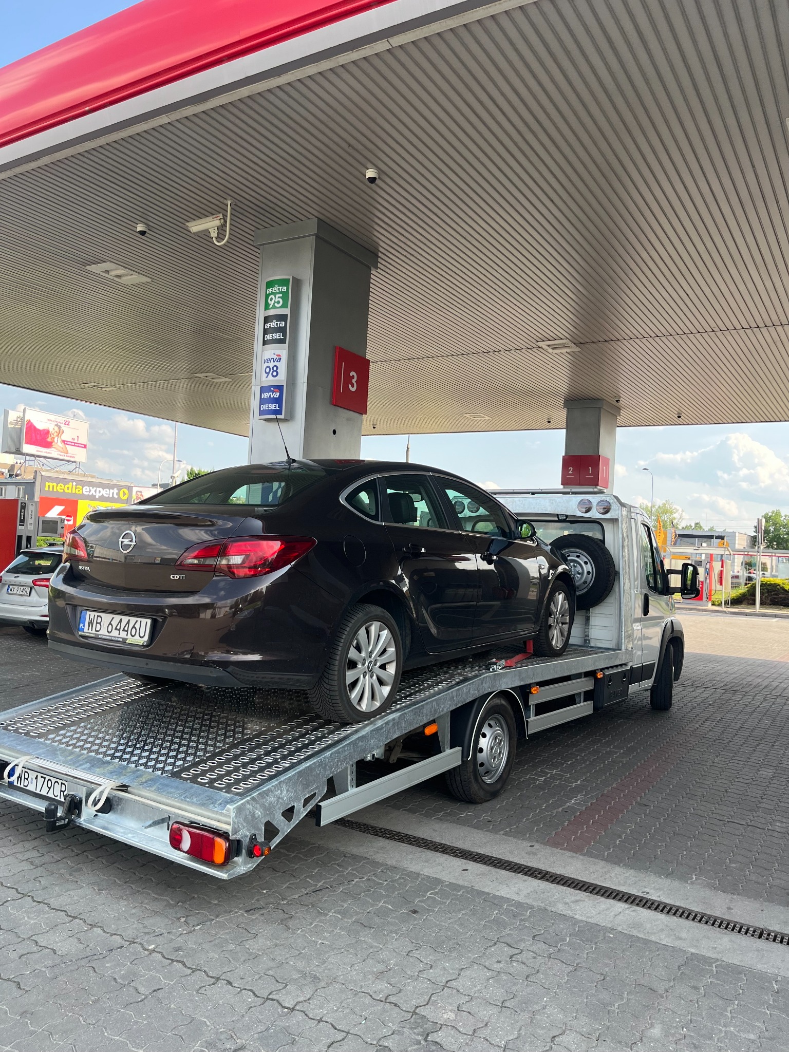 Brązowy Opel Astra na srebrnej platformie pomocy drogowej, na stacji benzynowej z dystrybutorami i cennikiem paliw w tle. Widoczne tablice rejestracyjne.