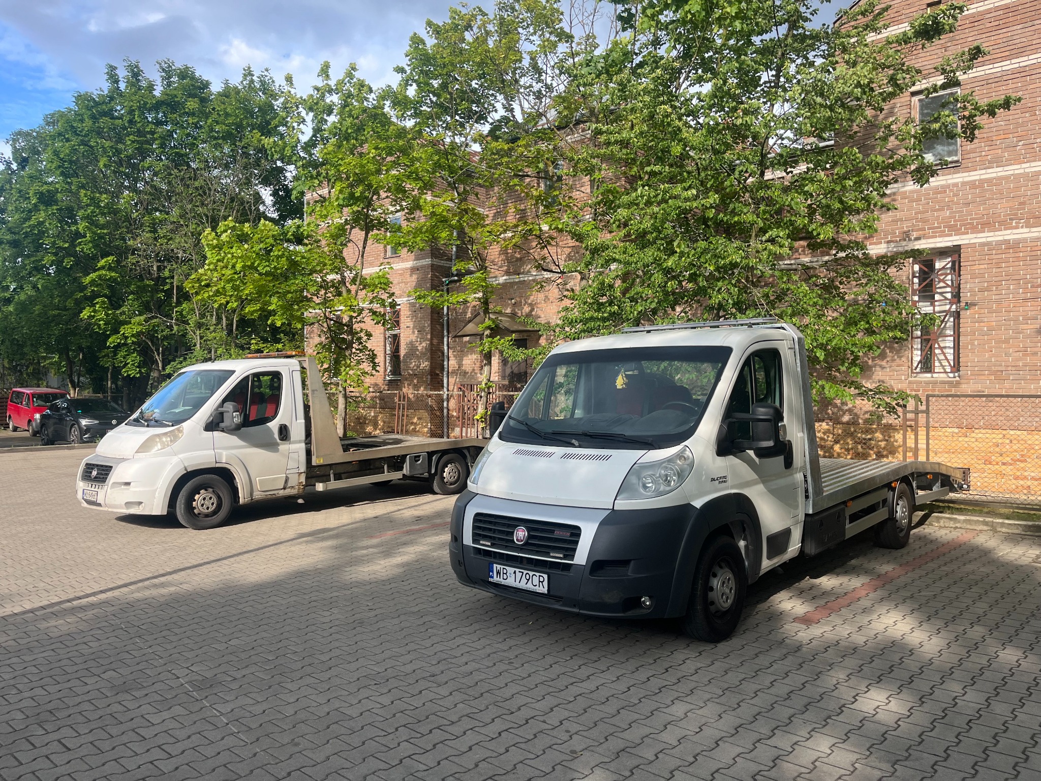Dwie białe lawety Fiat Ducato na parkingu z kostki brukowej, przed budynkiem z czerwonej cegły i zielonymi drzewami. Jedna laweta bliżej, druga w tle.