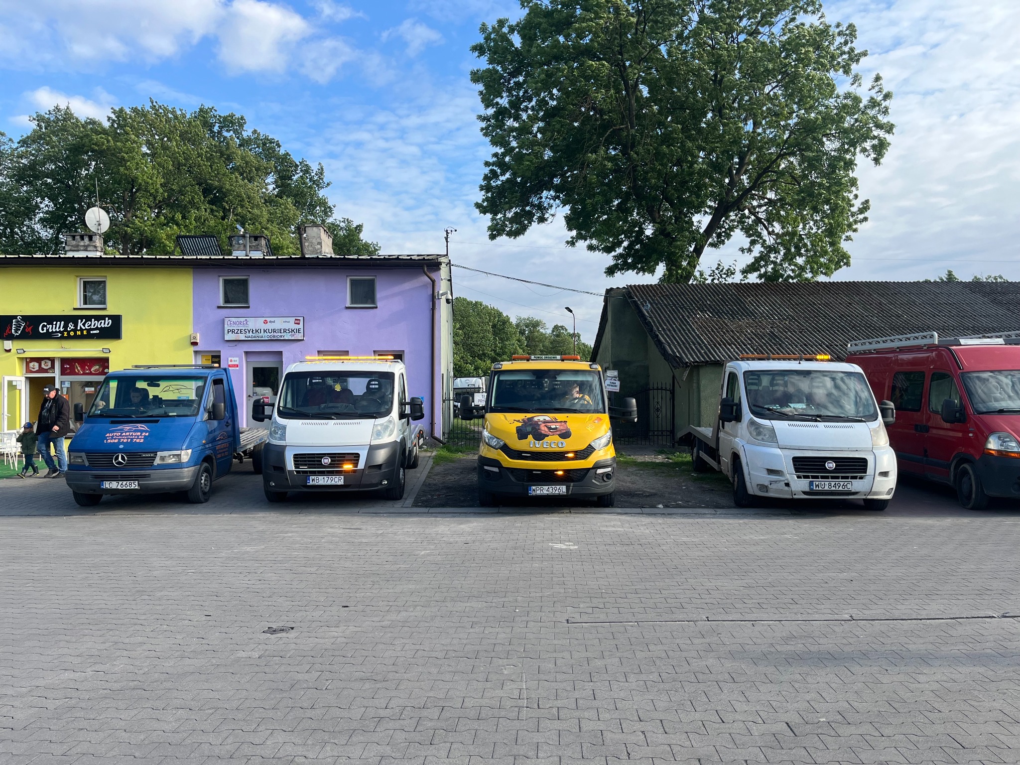 Flota pojazdów pomocy drogowej: niebieski Mercedes, biały Fiat, żółty Iveco z grafiką i czerwony Renault. Zaparkowane na brukowanym placu przed budynkami.