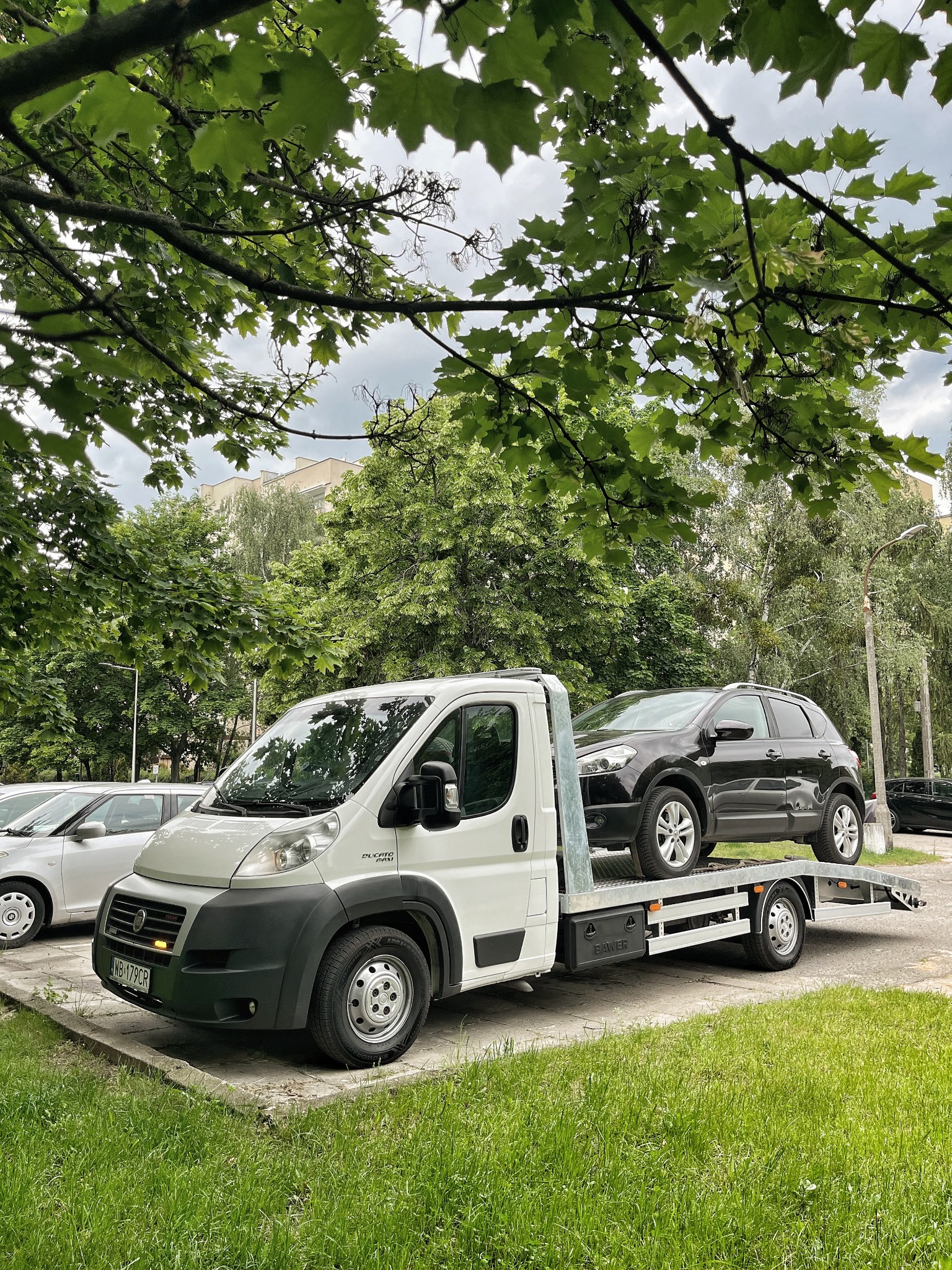 Laweta z czarnym SUV-em na platformie, zaparkowana na parkingu w otoczeniu zieleni miejskiej. Widoczne inne zaparkowane auta i budynek w tle.
