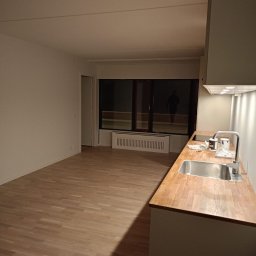 GoldenHand - Wykończona kuchnia z drewnianym blatem i parkietem. W tle okno z widoczną osobą na zewnątrz. Świeżo po remoncie, gotowa do użytku. Nowoczesny minimalizm.