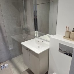 GoldenHand - Nowoczesna łazienka po remoncie: szare płytki, kabina prysznicowa, podwieszana umywalka z szafką i toaleta. Na podłodze widoczny odkurzacz budowlany.