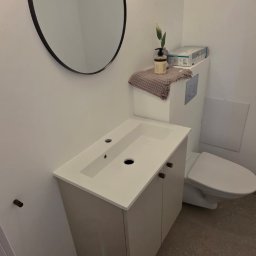 GoldenHand - Nowoczesna łazienka z okrągłym lustrem nad białą szafką umywalkową i toaletą. Stonowana kolorystyka, minimalistyczny design, oświetlenie punktowe.