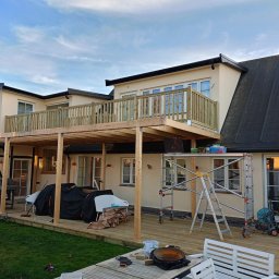 GoldenHand - Drewniana konstrukcja tarasu z balustradą w Zgierzu, częściowo zadaszona, w trakcie budowy przy beżowym budynku. Widoczne rusztowania i narzędzia.