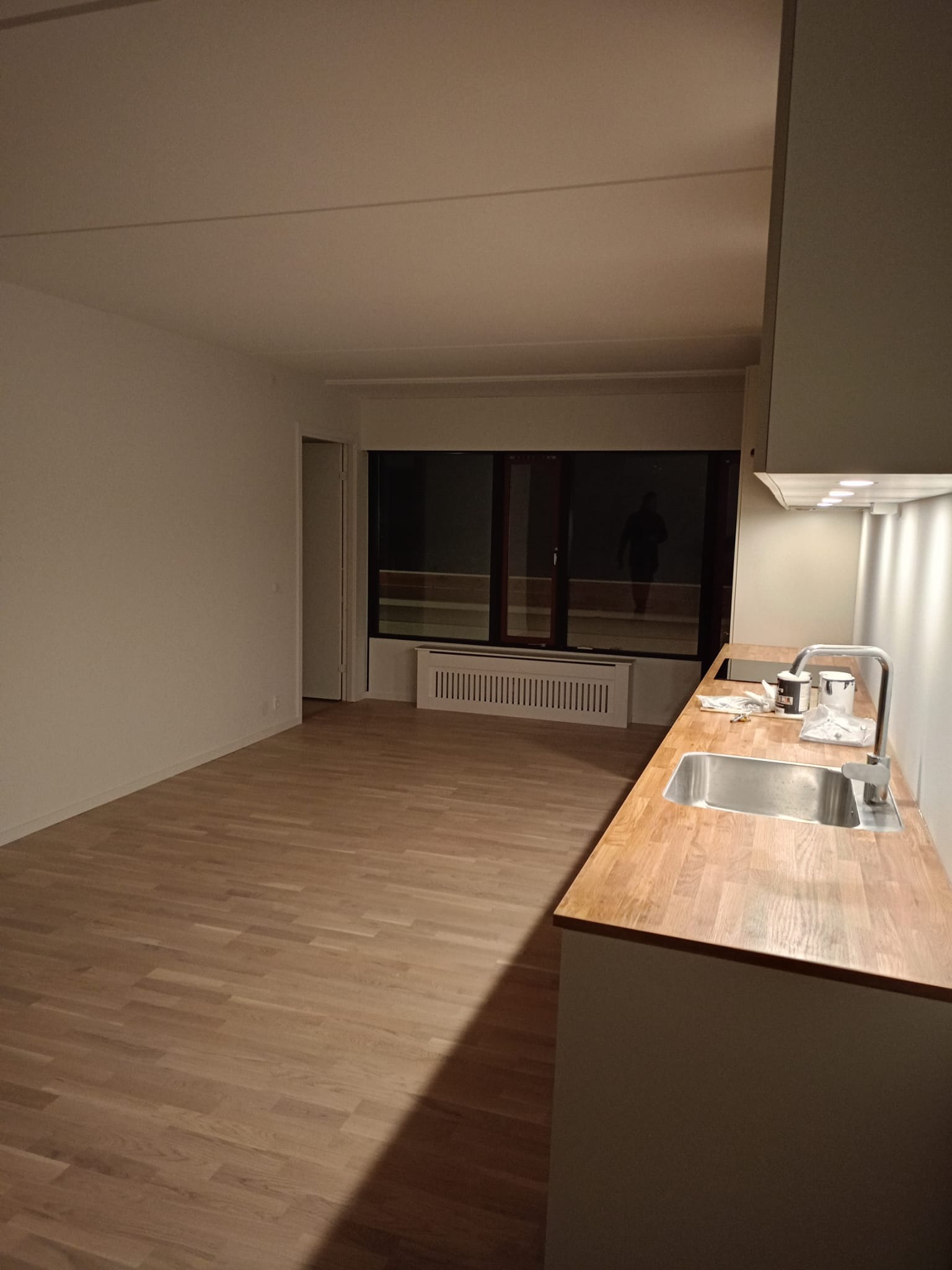 Wykończona kuchnia z drewnianym blatem i parkietem. W tle okno z widoczną osobą na zewnątrz. Świeżo po remoncie, gotowa do użytku. Nowoczesny minimalizm.