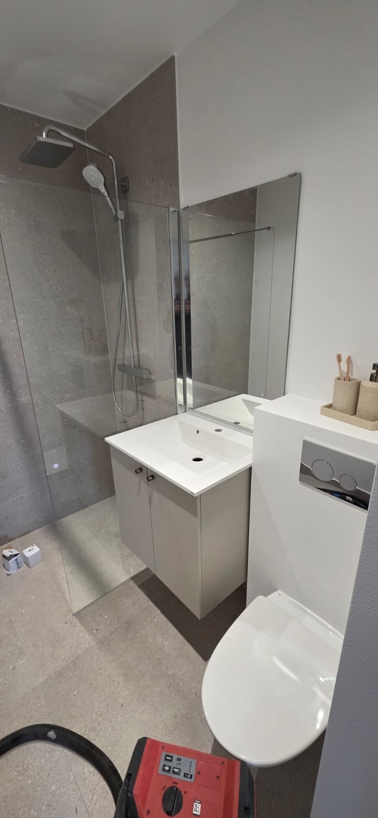 Nowoczesna łazienka po remoncie: szare płytki, kabina prysznicowa, podwieszana umywalka z szafką i toaleta. Na podłodze widoczny odkurzacz budowlany.