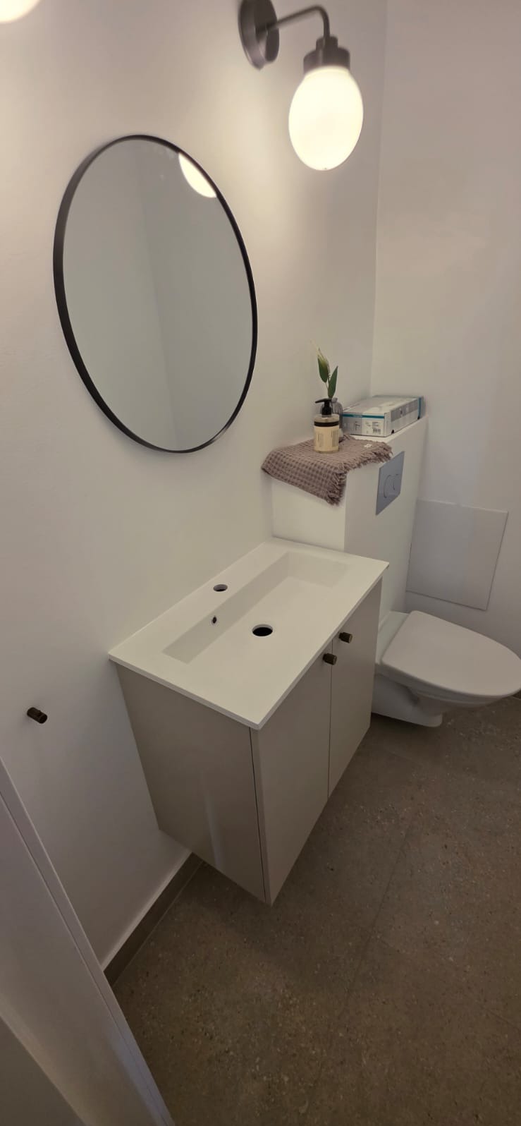 Nowoczesna łazienka z okrągłym lustrem nad białą szafką umywalkową i toaletą. Stonowana kolorystyka, minimalistyczny design, oświetlenie punktowe.