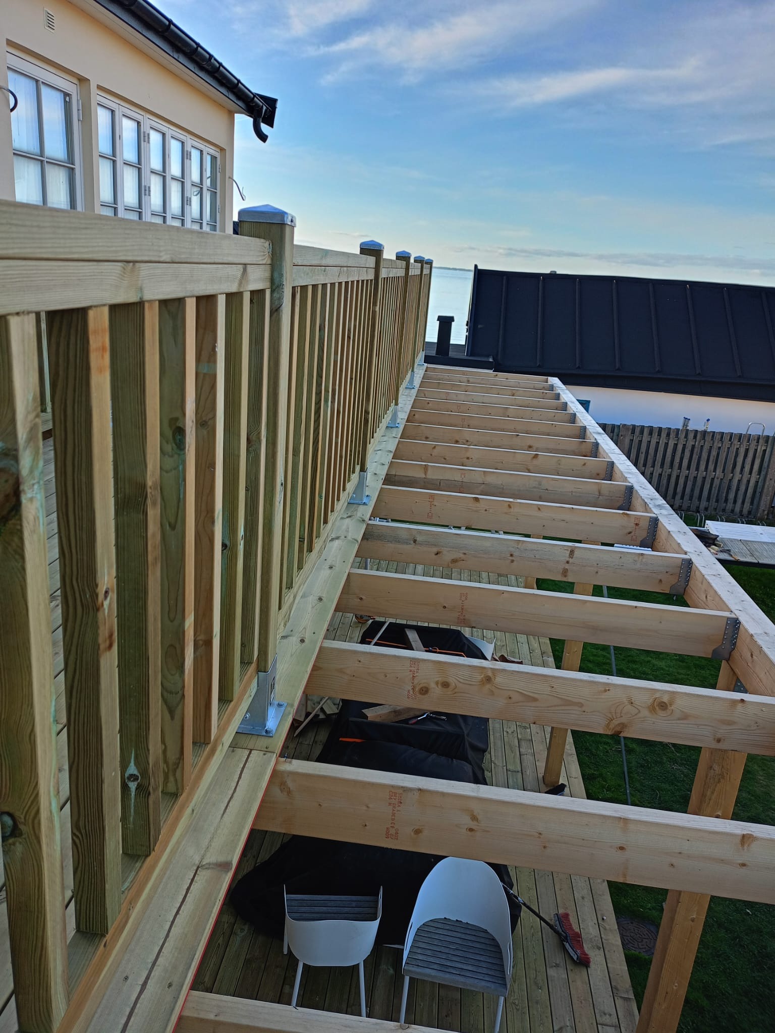 Drewniana konstrukcja tarasu w trakcie budowy, widok z góry. W tle dom z oknami i fragment nieba. Jasne drewno, metalowe łączniki, częściowo ułożona podłoga.