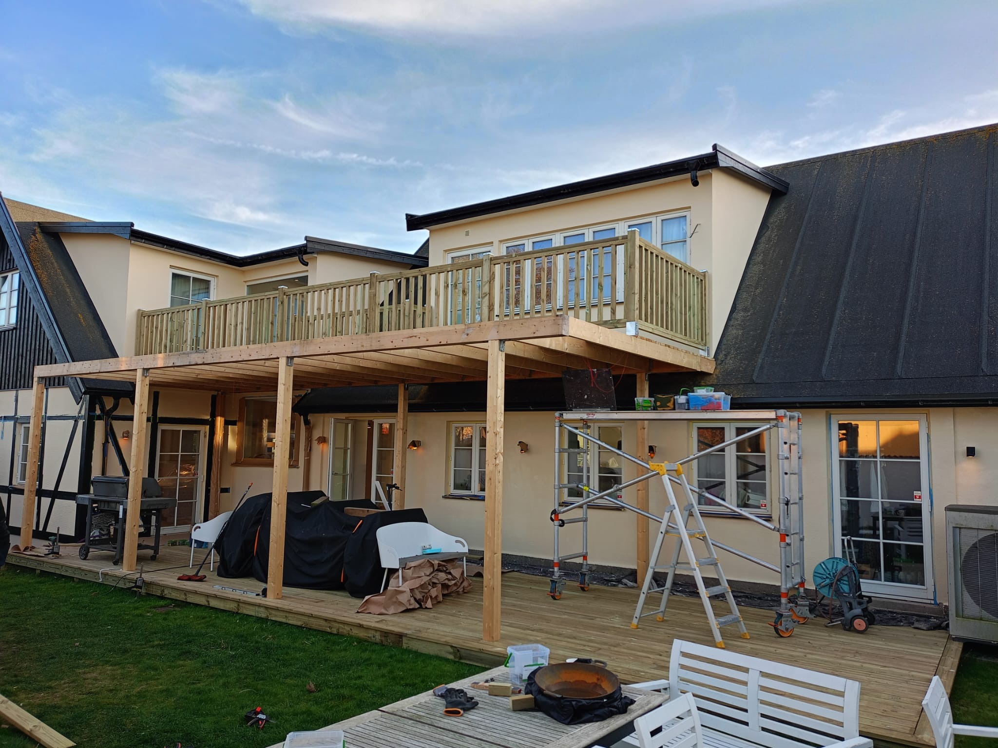 Drewniana konstrukcja tarasu z balustradą w Zgierzu, częściowo zadaszona, w trakcie budowy przy beżowym budynku. Widoczne rusztowania i narzędzia.