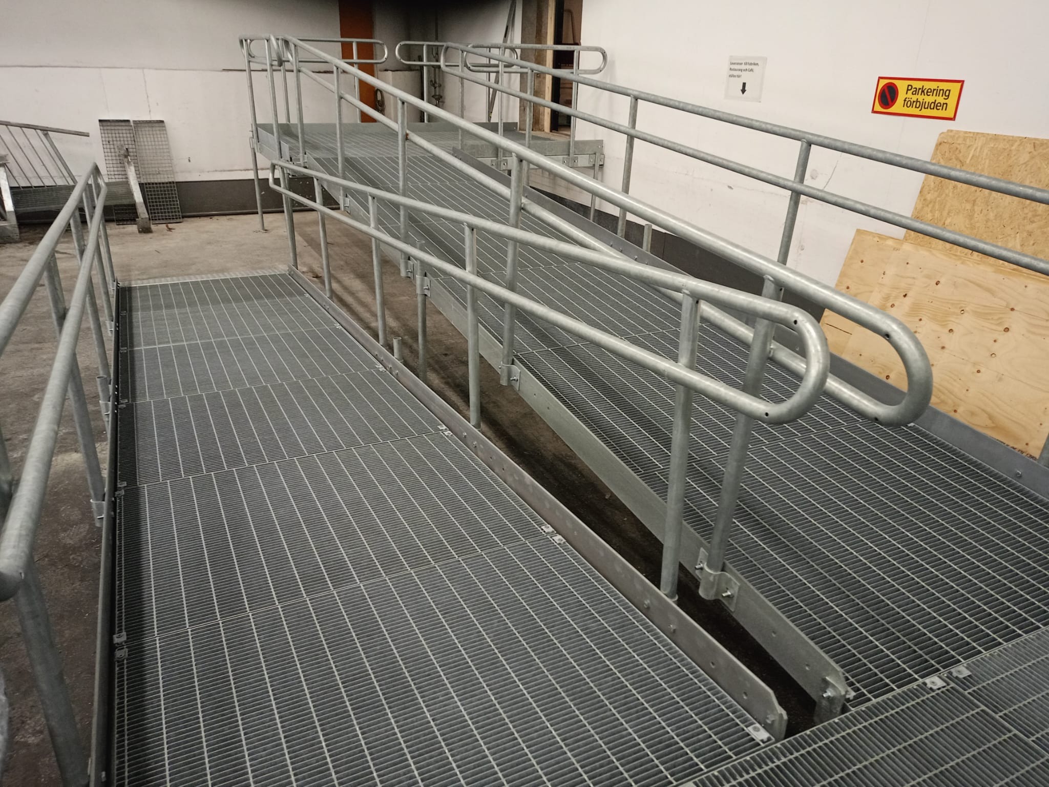 Metalowa rampa dla wózków inwalidzkich z poręczami, z kratką na podłodze, w szarym kolorze, wewnątrz budynku, z tabliczką informacyjną na ścianie. Ujęcie z dołu.