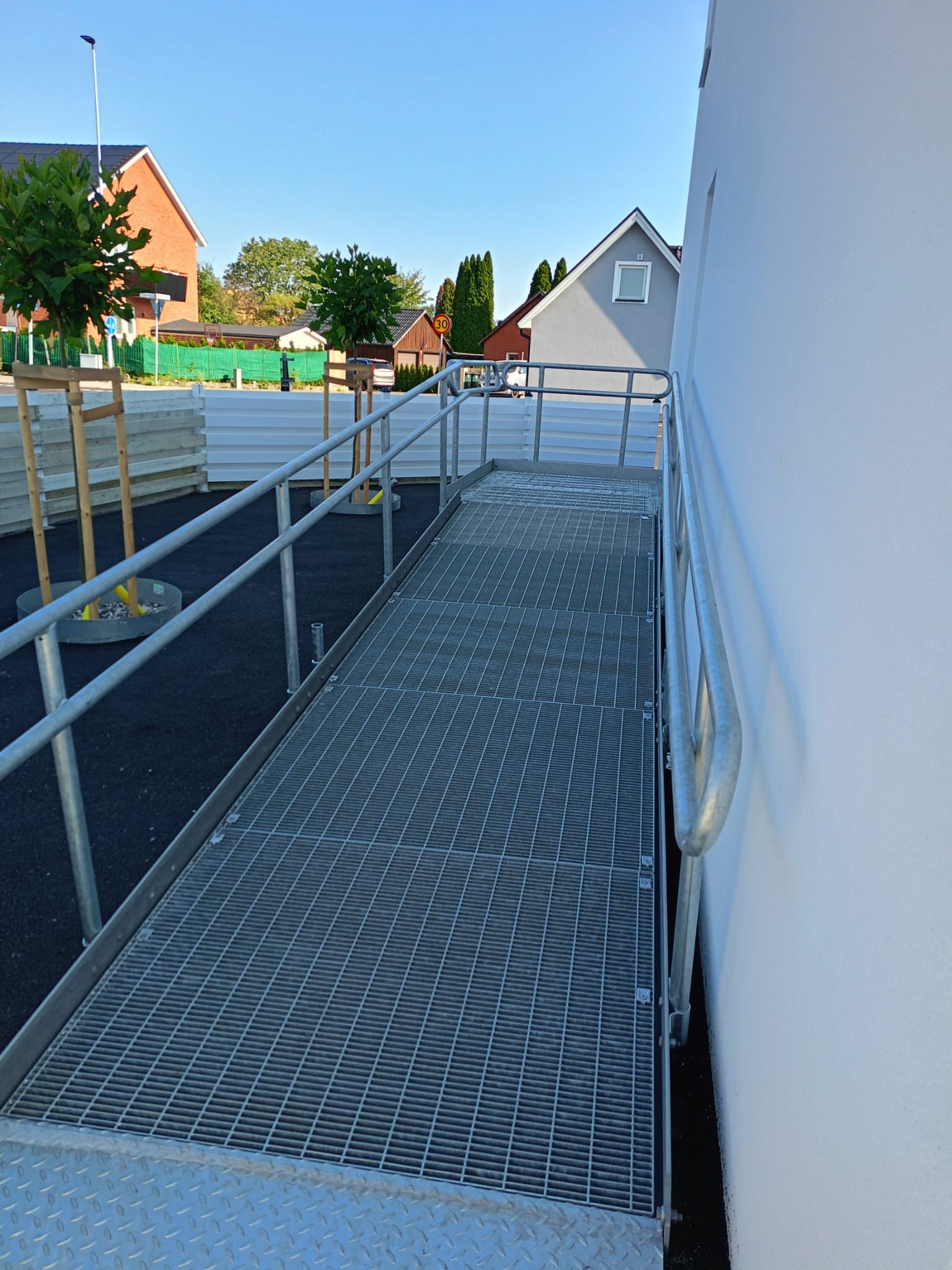 Zewnętrzna, metalowa rampa z poręczami, prowadząca do budynku z białą elewacją. W tle domy jednorodzinne i zieleń. Widok w pogodny dzień.