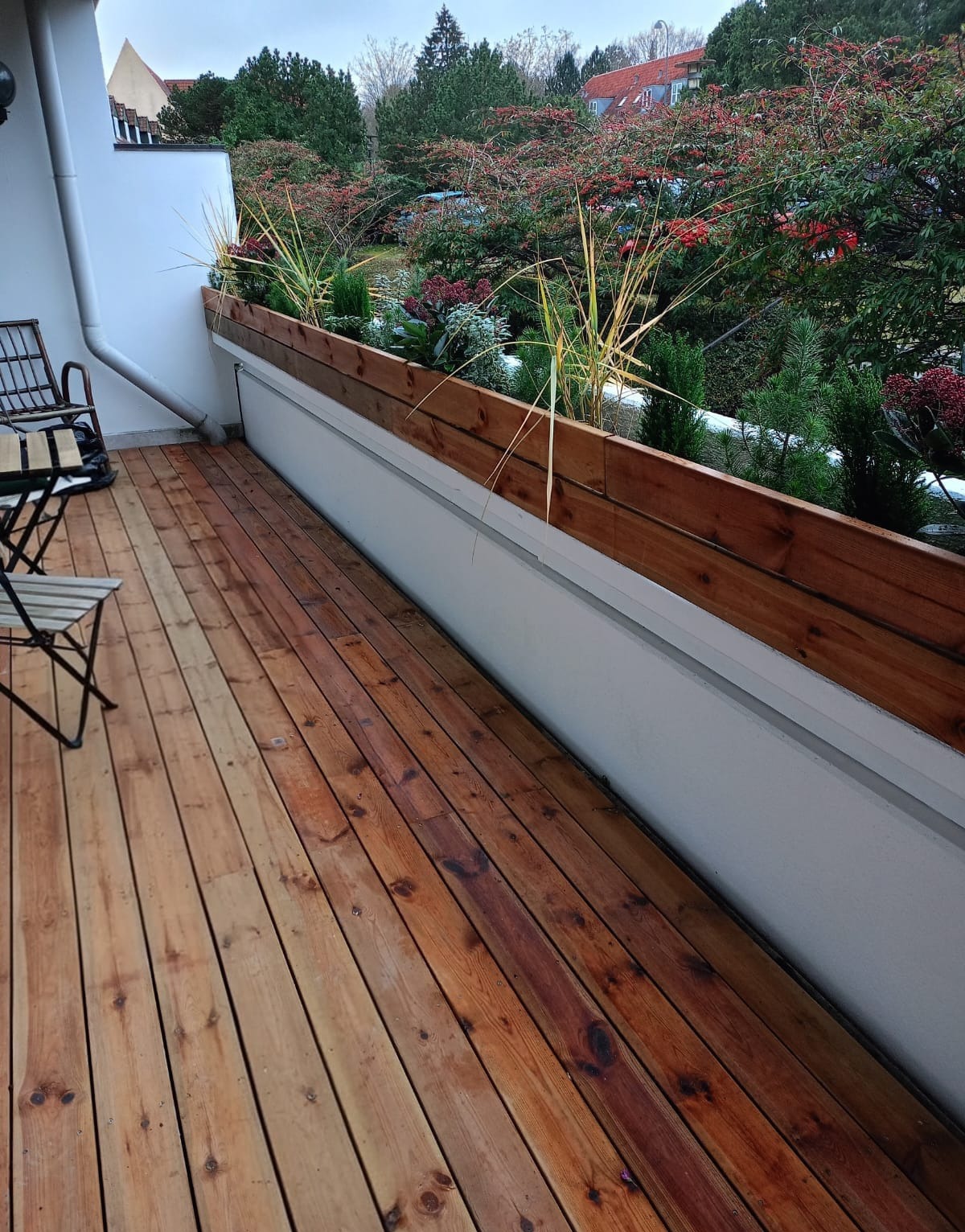 Drewniany balkon z roślinami ozdobnymi w donicach, widok z góry na deski podłogowe i balustradę. W tle zieleń drzew i krzewów.