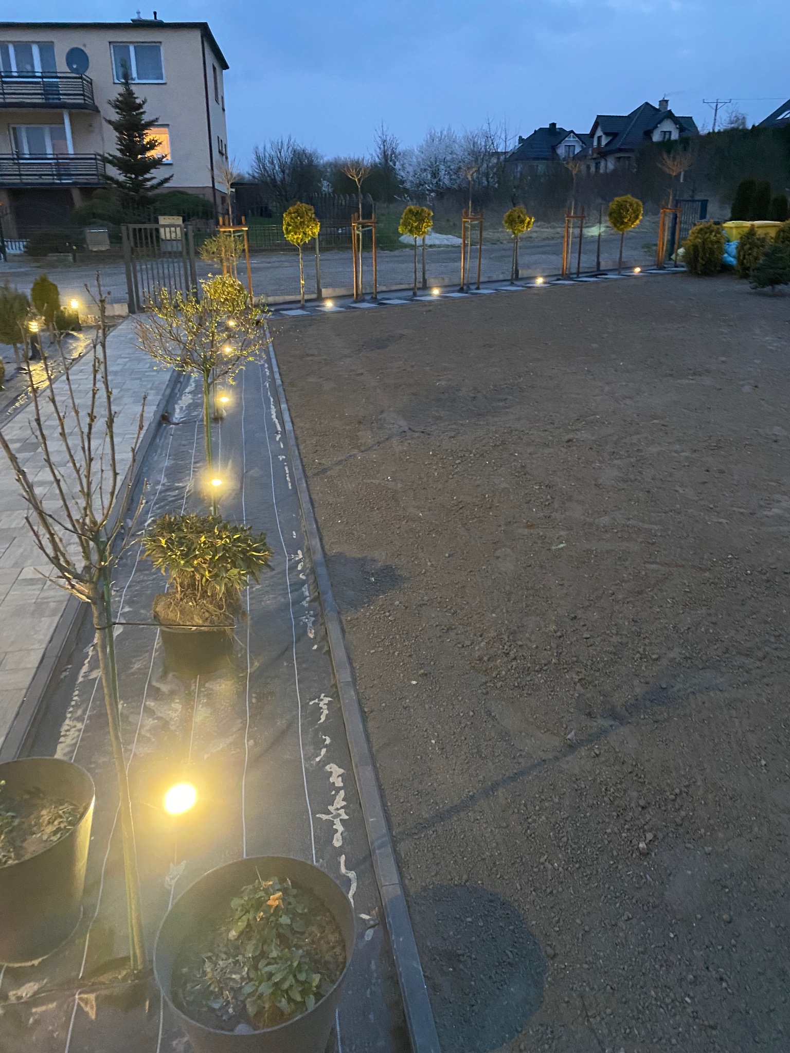 Przygotowanie terenu pod trawnik z nasadzeniami: drzewka w donicach, geowłóknina, obrzeża i oświetlenie wzdłuż ścieżki. W tle budynek mieszkalny i linia drzew.