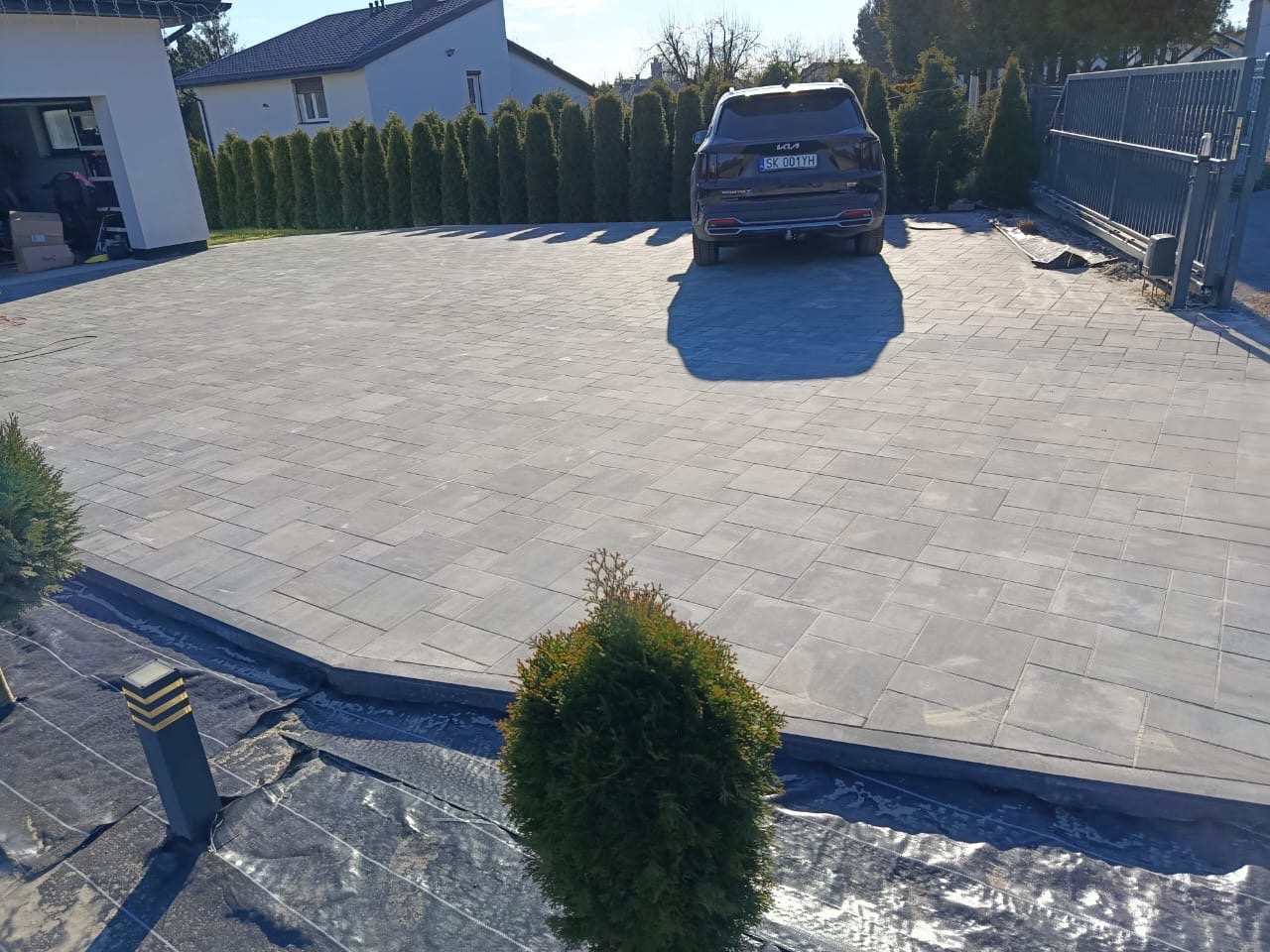 Szara kostka brukowa na podjeździe domu jednorodzinnego, widok z góry, czarny samochód zaparkowany na końcu podjazdu, tuje w tle, lampy oświetleniowe wzdłuż krawędzi.