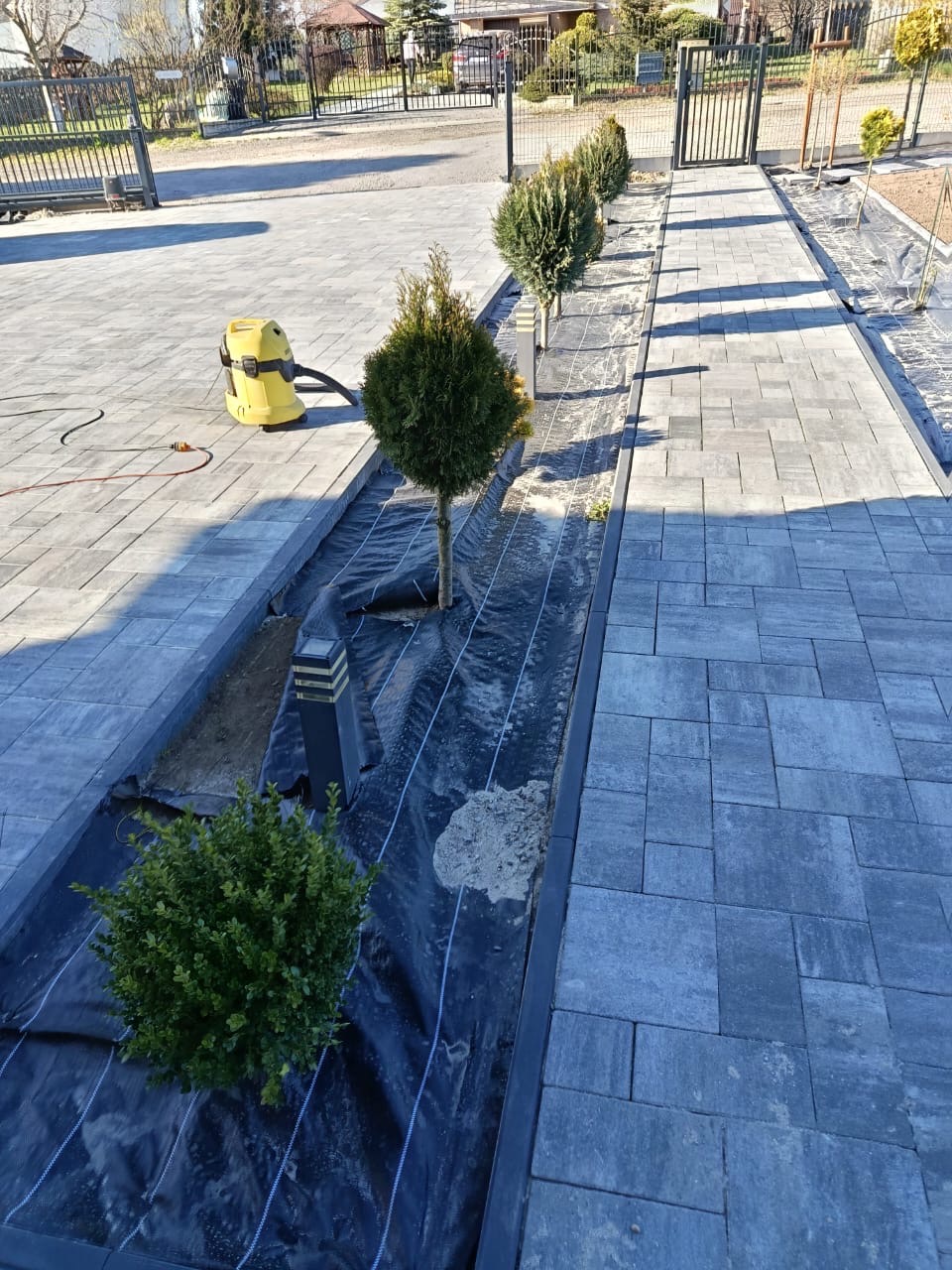 Ułożona szara kostka brukowa z wykończonym obrzeżem rabaty, w której posadzono rośliny. Na kostce stoi żółty odkurzacz przemysłowy. Widok na metalową bramę w tle.