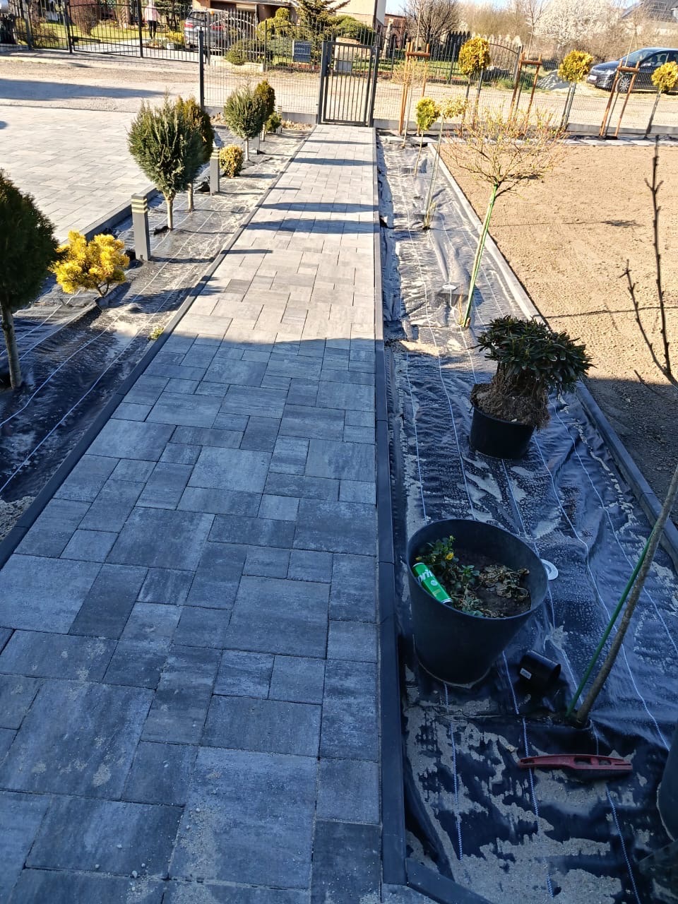 Ułożona ścieżka z kostki brukowej prowadząca do bramy. Po prawej stronie przygotowane podłoże pod nasadzenia, rozłożona agrowłóknina, donice z roślinami.