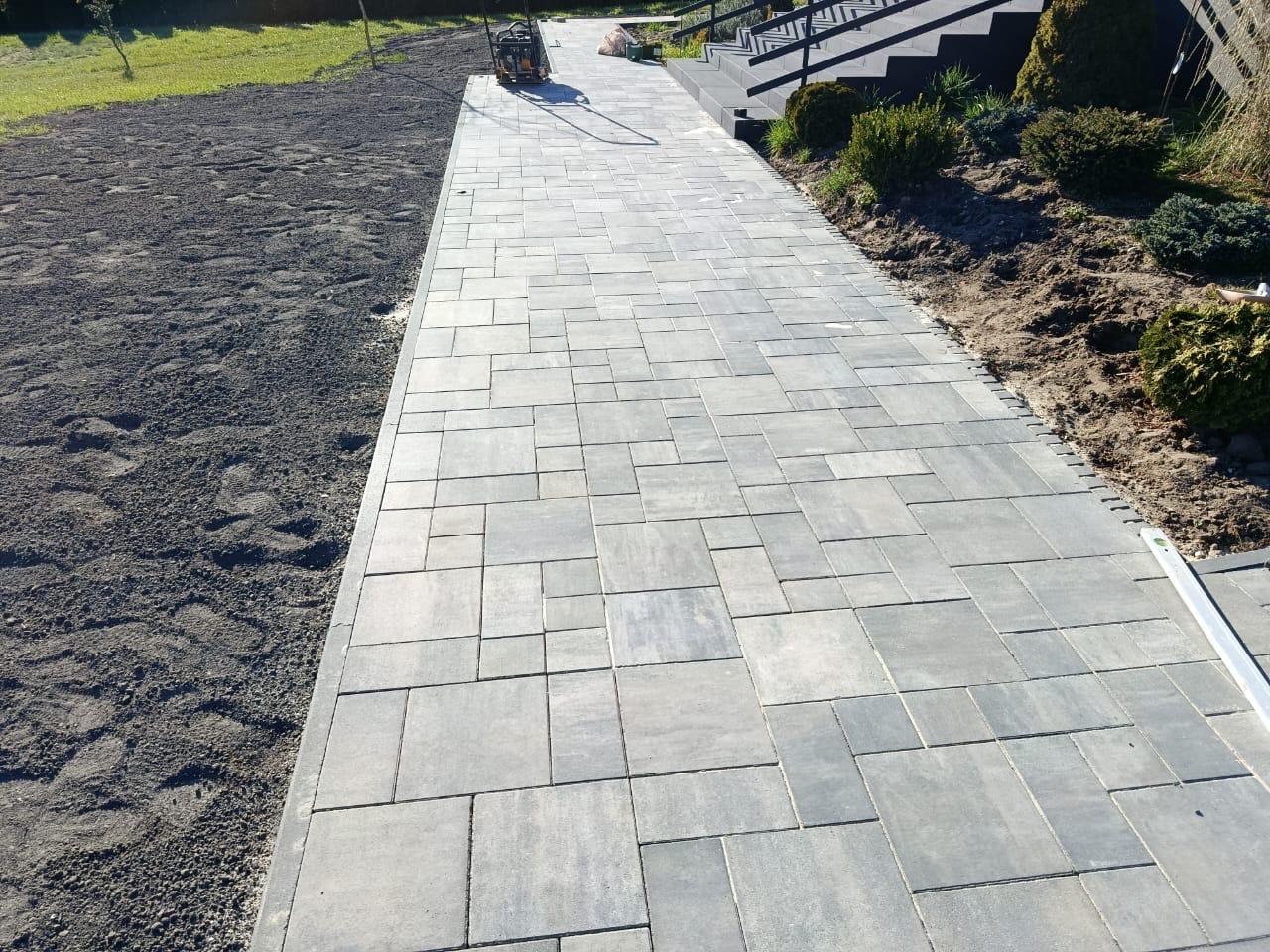 Ułożona ścieżka z szarej kostki brukowej o nieregularnym wzorze, prowadząca od trawnika do schodów wejściowych, z widoczną ubijarką w tle i elementami krajobrazu.