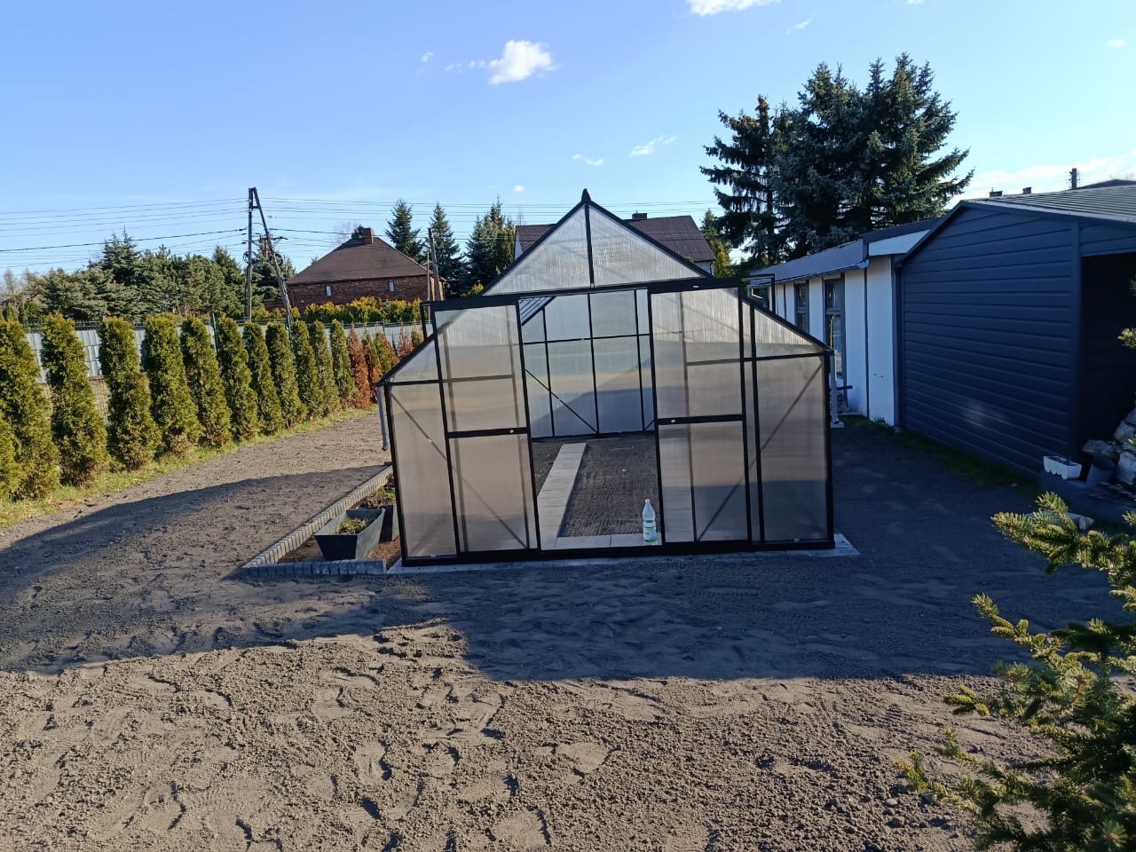 Nowoczesna szklarnia z czarną ramą, otoczona przygotowanym podłożem, w tle zielone tuje i budynki. Słoneczny dzień.