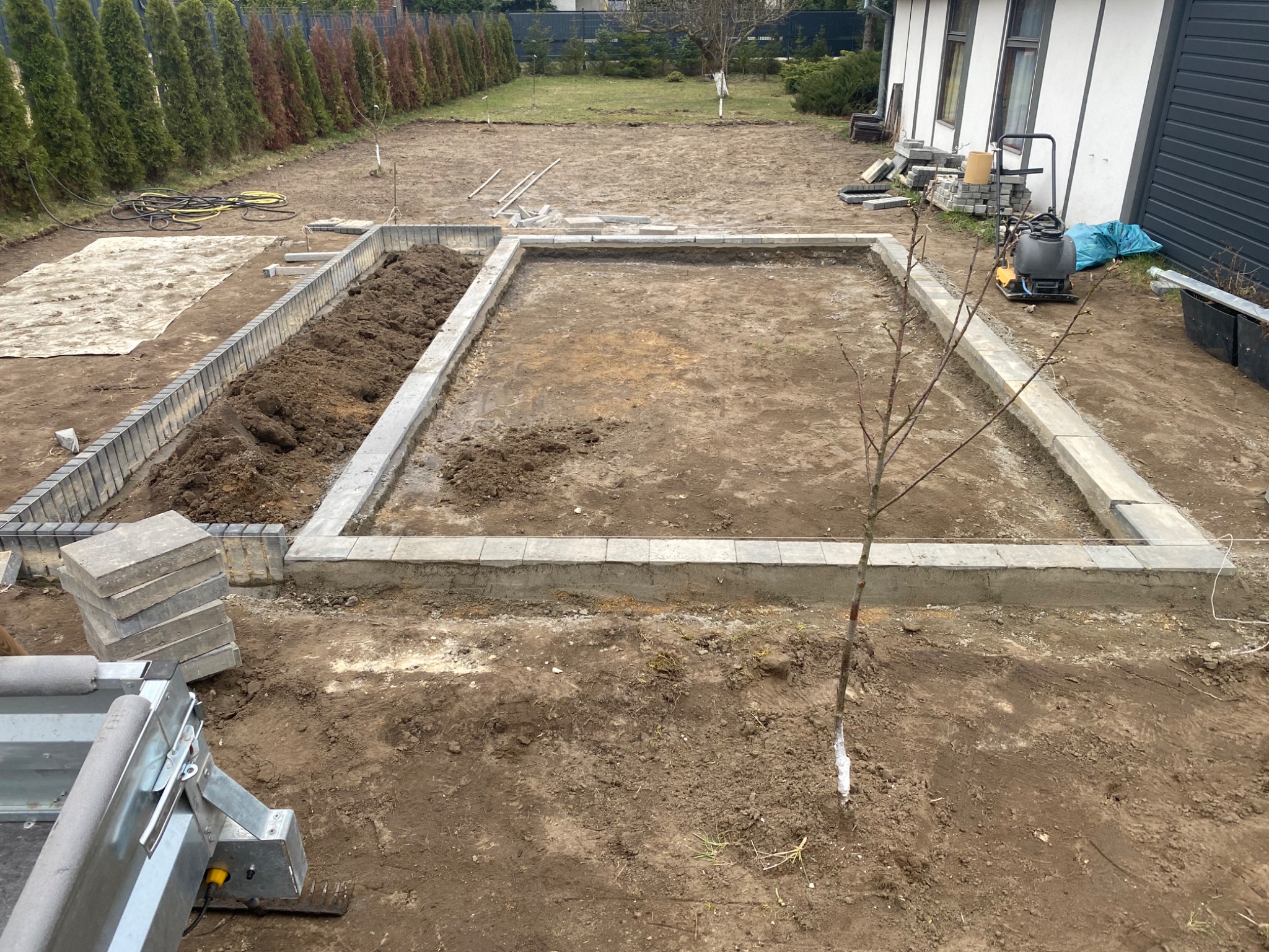 Etap prac: obrzeża z kostki betonowej, częściowo wypełnione ziemią, tworzą ramę na posesji. W tle rząd tuj i budynek. Widoczny sprzęt budowlany.