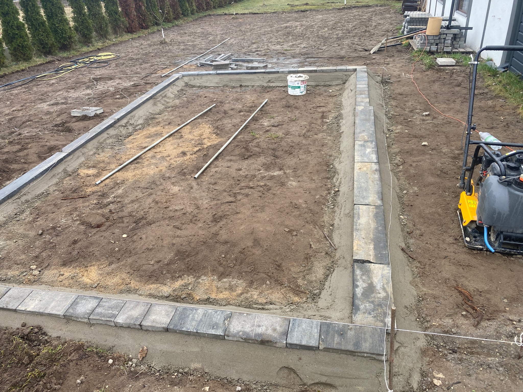 Wyrównany teren pod budowę, obrzeża z kostki, zagęszczarka w tle. Widoczne narzędzia i materiały budowlane. Etap przygotowania podłoża.