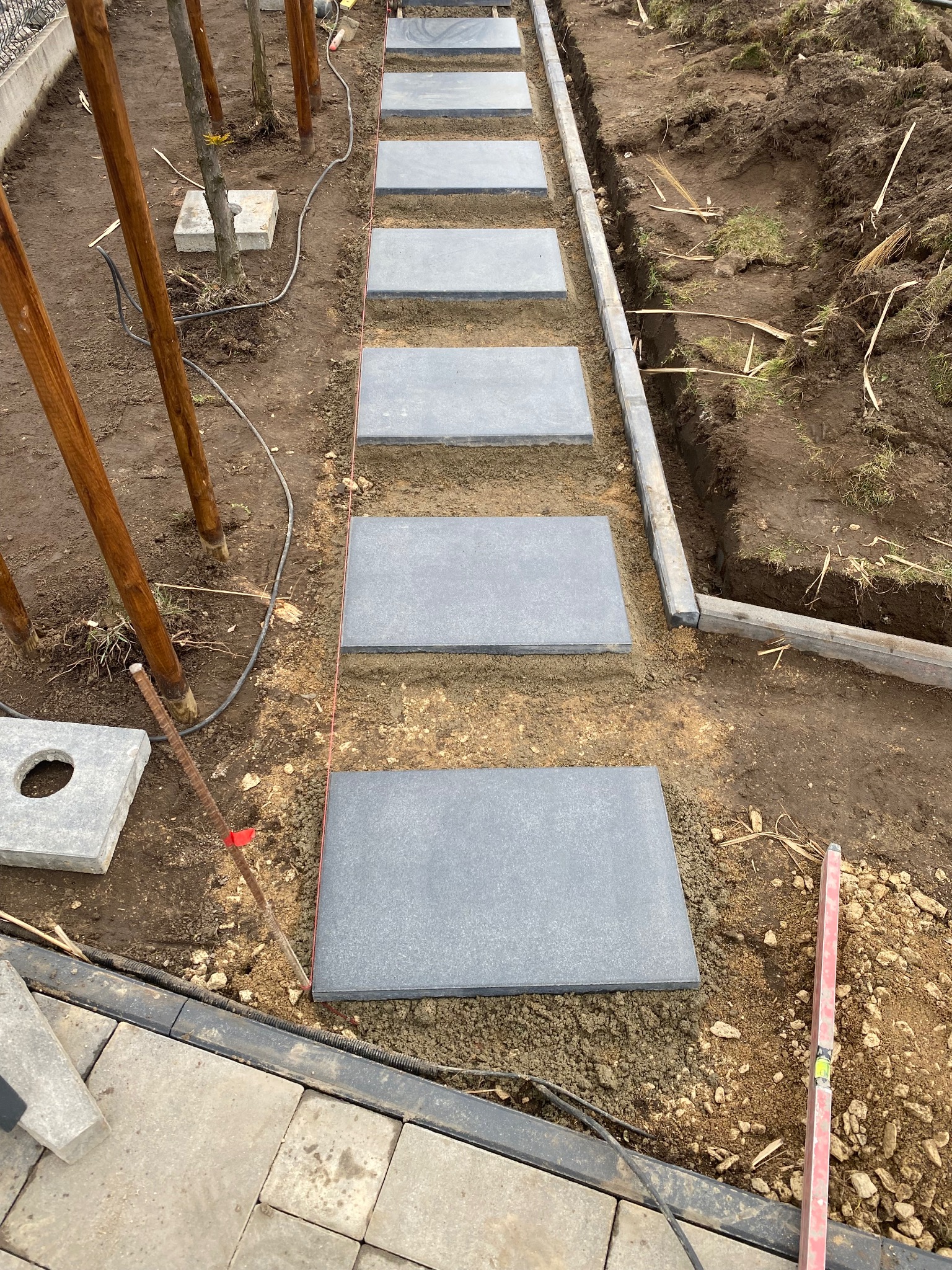 Układanie ścieżki z szarych płyt na gruncie, widoczne drewniane słupki, poziomica i sznurek wyznaczający linię. Etap prac brukarskich w ogrodzie.