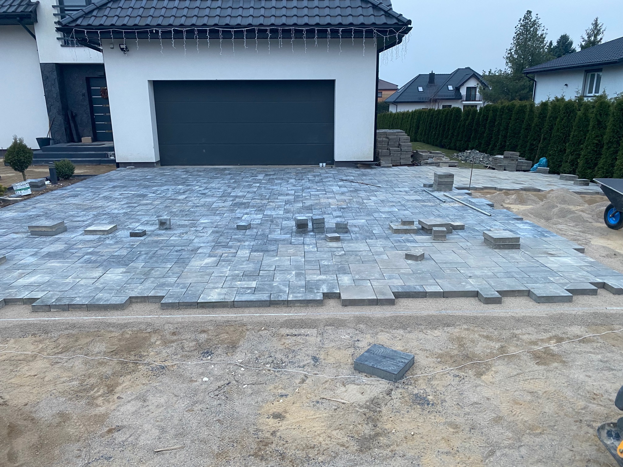 W trakcie układania szarej kostki brukowej na podjeździe przed nowoczesnym domem z grafitową bramą garażową. Częściowo ułożona nawierzchnia, widoczne narzędzia i materiały.