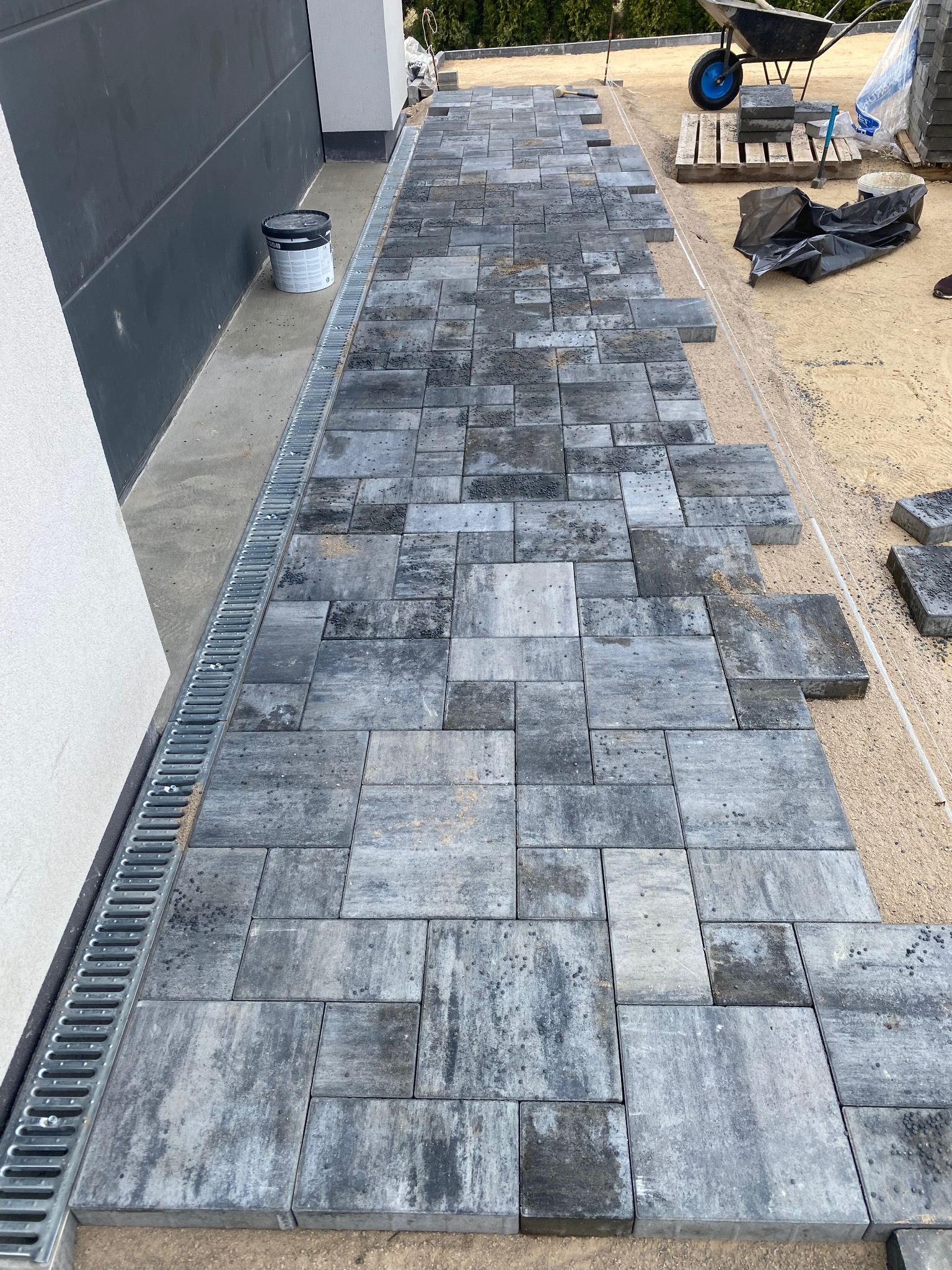 Fragment układanej ścieżki z szarej kostki brukowej, widoczny drenaż liniowy przy budynku, taczka i paleta z kostką w tle. Etap prac brukarskich.