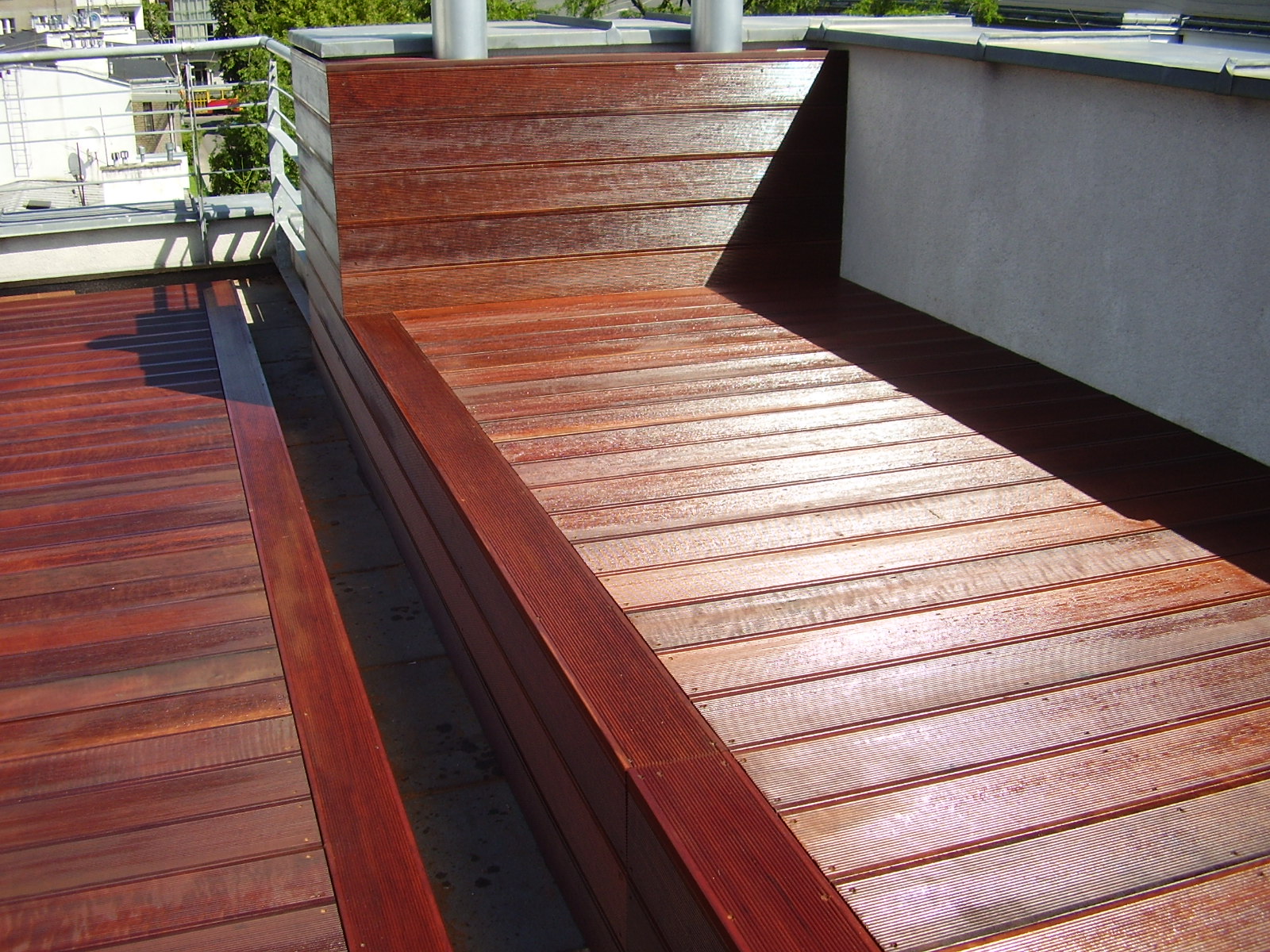 Czerwono-brązowy taras z desek kompozytowych na dachu budynku, z wbudowaną ławką i metalową balustradą, widziany z góry, w słoneczny dzień.
