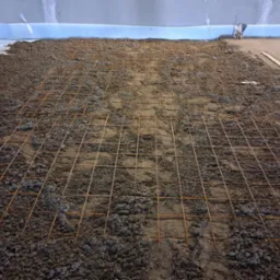 Przygotowanie podłoża pod posadzkę: siatka zbrojeniowa na warstwie zaprawy. Widoczna izolacja przy ścianie i drewniana listwa. Ujęcie z góry, perspektywa zbieżna.