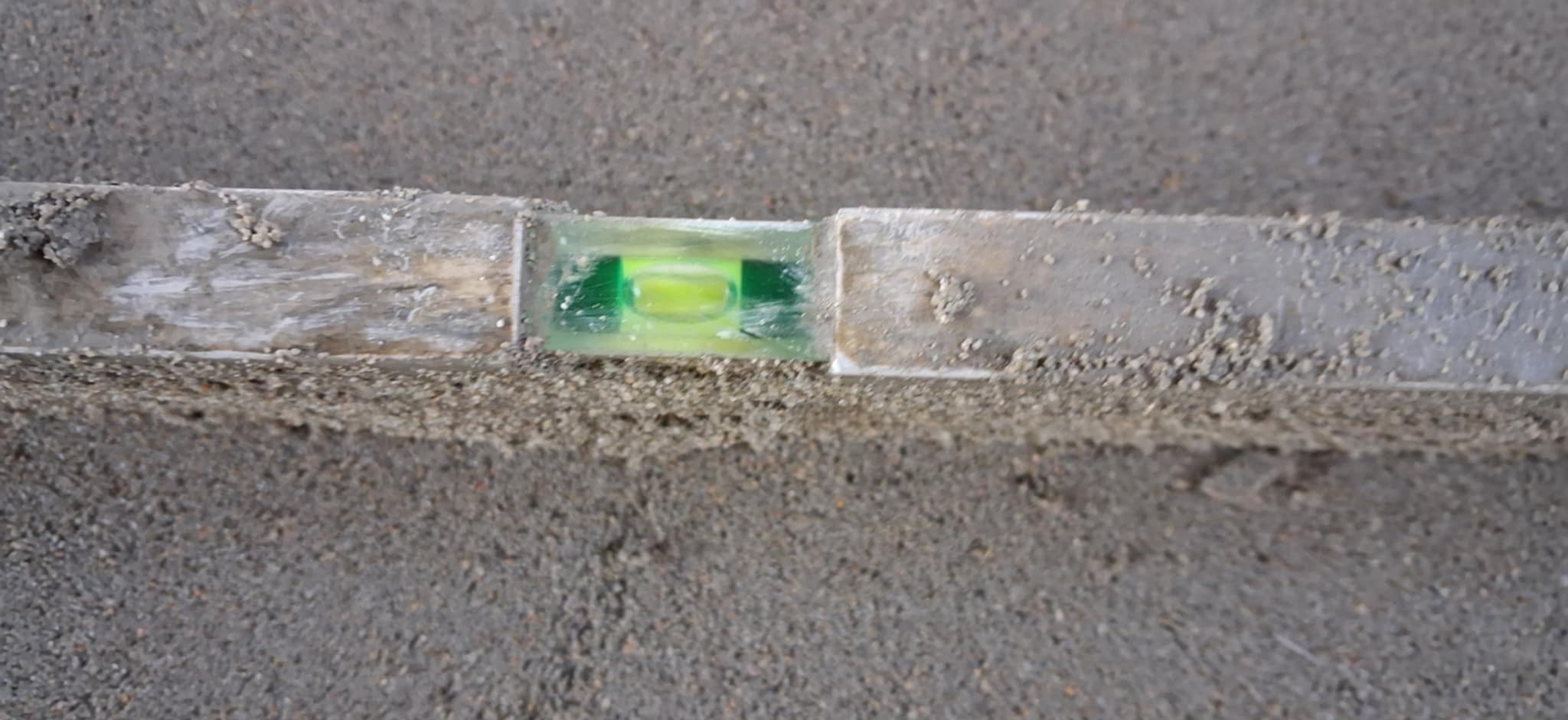 Poziomnica na szarej, surowej posadzce jastrychowej, z bliska. Widoczny pęcherzyk powietrza w zielonym płynie. Szorstka powierzchnia, detale wykończenia.