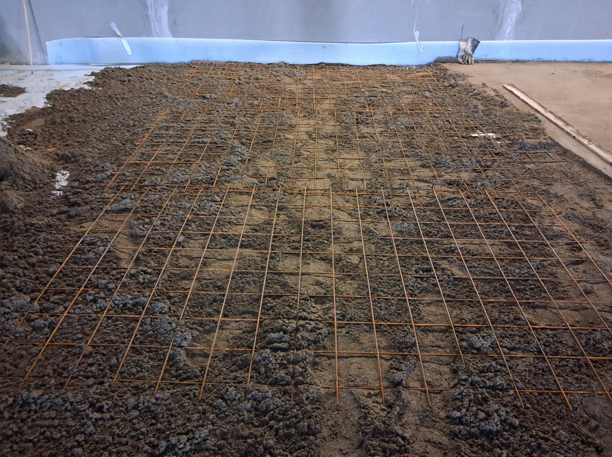 Przygotowanie podłoża pod posadzkę: siatka zbrojeniowa na warstwie zaprawy. Widoczna izolacja przy ścianie i drewniana listwa. Ujęcie z góry, perspektywa zbieżna.