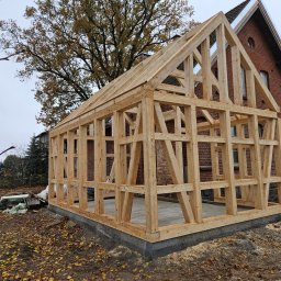 Michał Zaworski domy drewniane. - Konstrukcja domu szkieletowego w trakcie budowy, widoczny drewniany szkielet na fundamencie, w tle budynek z czerwonej cegły i drzewo.