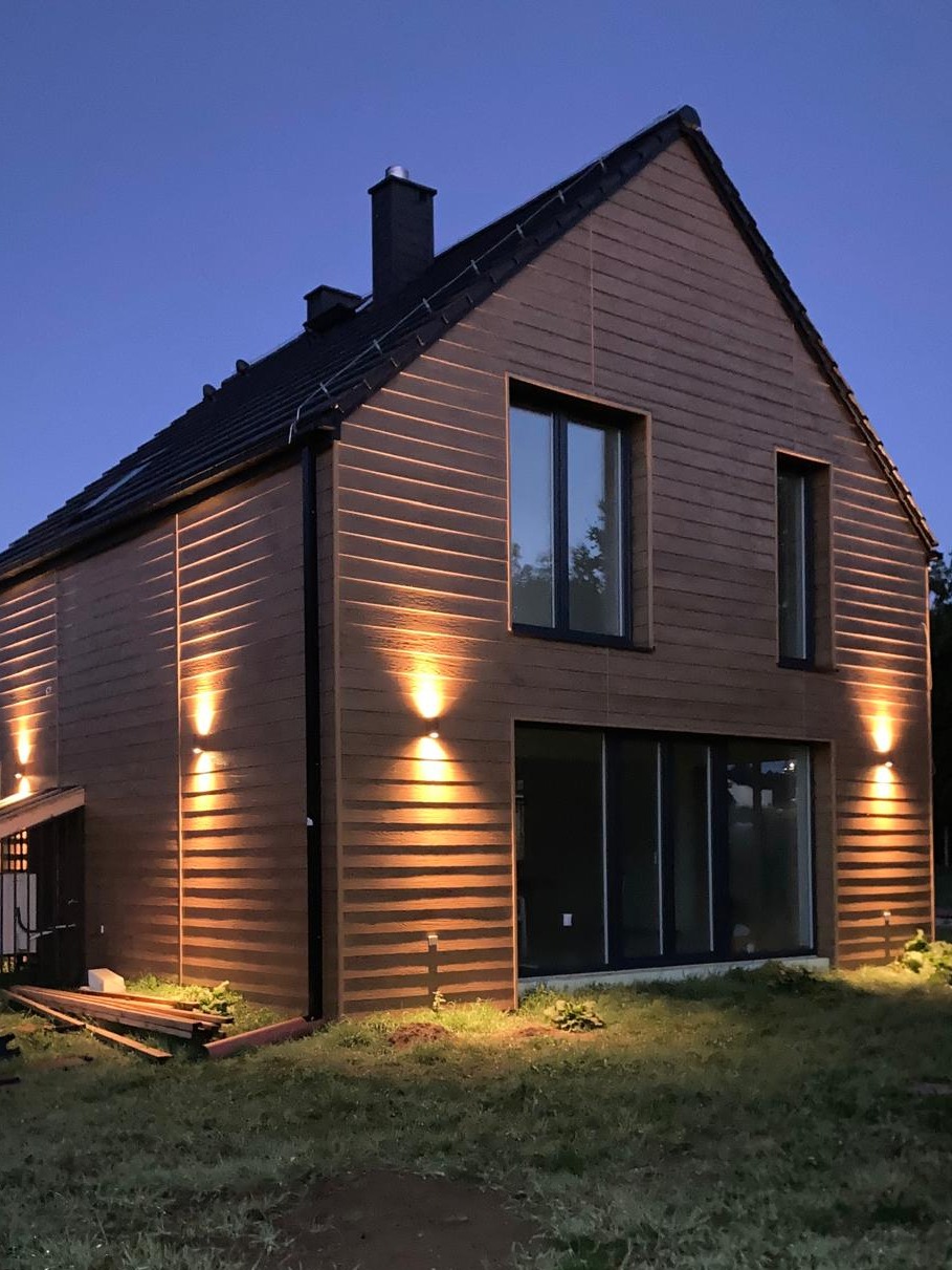 Nowoczesny dom z drewnianą elewacją i oświetleniem zewnętrznym, sfotografowany o zmierzchu. Widoczne duże okna i komin.