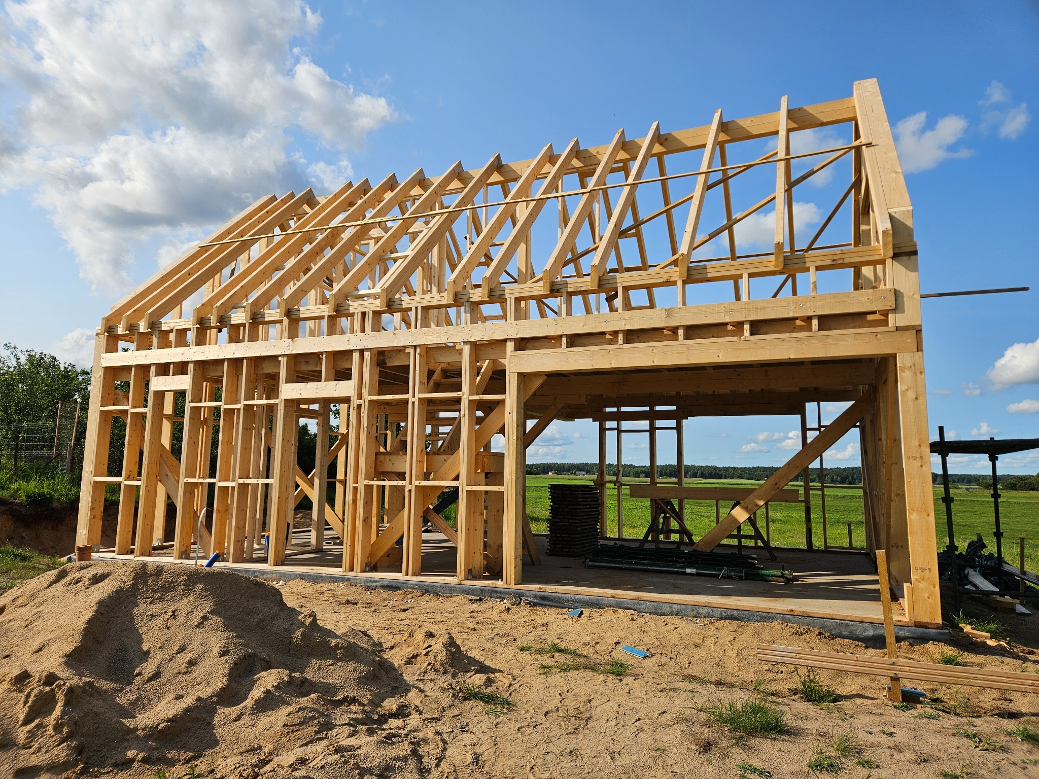Wznoszona konstrukcja szkieletowa domu z jasnego drewna na fundamencie, z widocznym stosem materiałów budowlanych w tle i błękitnym niebem.