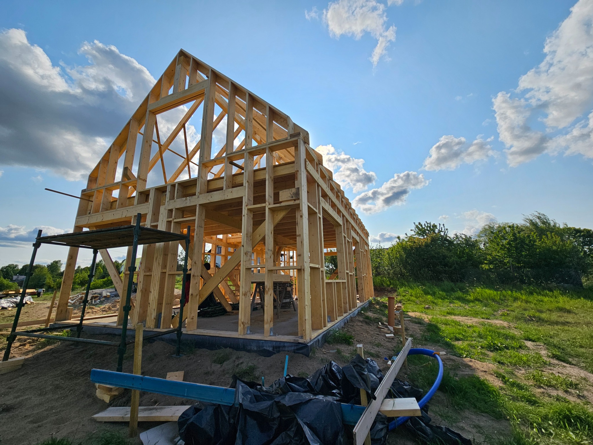 Konstrukcja drewniana domu w trakcie budowy, widok z dołu na szkielet budynku pod błękitnym niebem z chmurami. Widoczne rusztowanie i teren budowy.