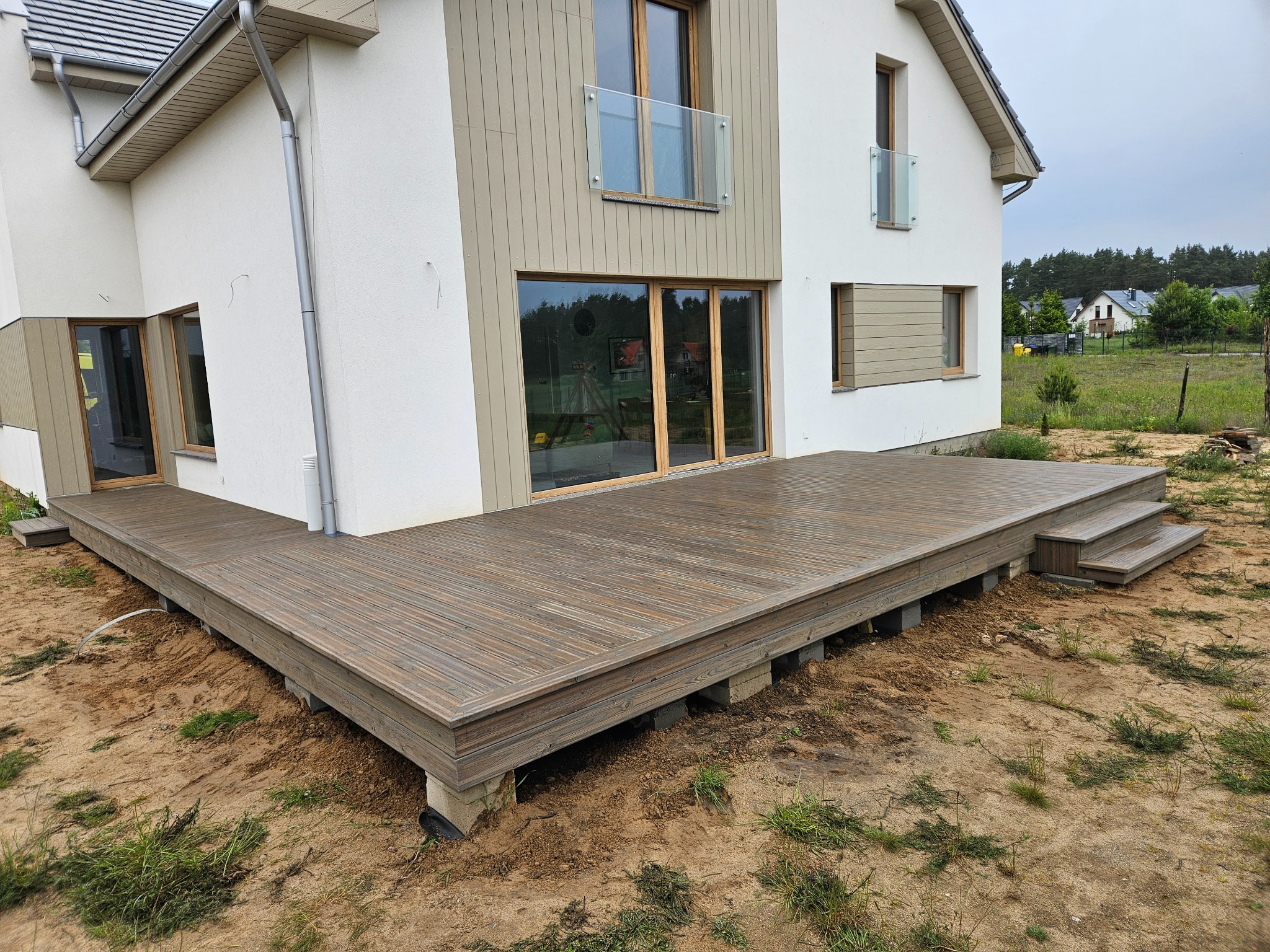 Drewniany taras przy nowoczesnym domu szkieletowym z białymi ścianami i drewnianą elewacją. Widok na taras z niskiej perspektywy w pochmurny dzień.